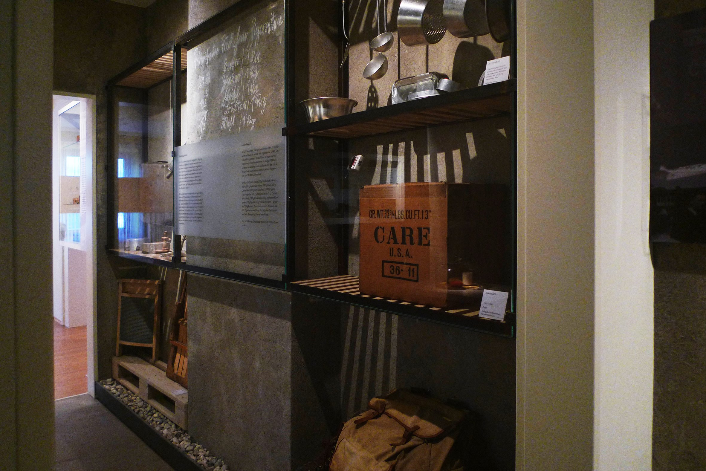 Innenansicht eines Museums mit Glaskästen, die Gegenstände wie einen Karton, Werkzeug und Geschirr enthalten.