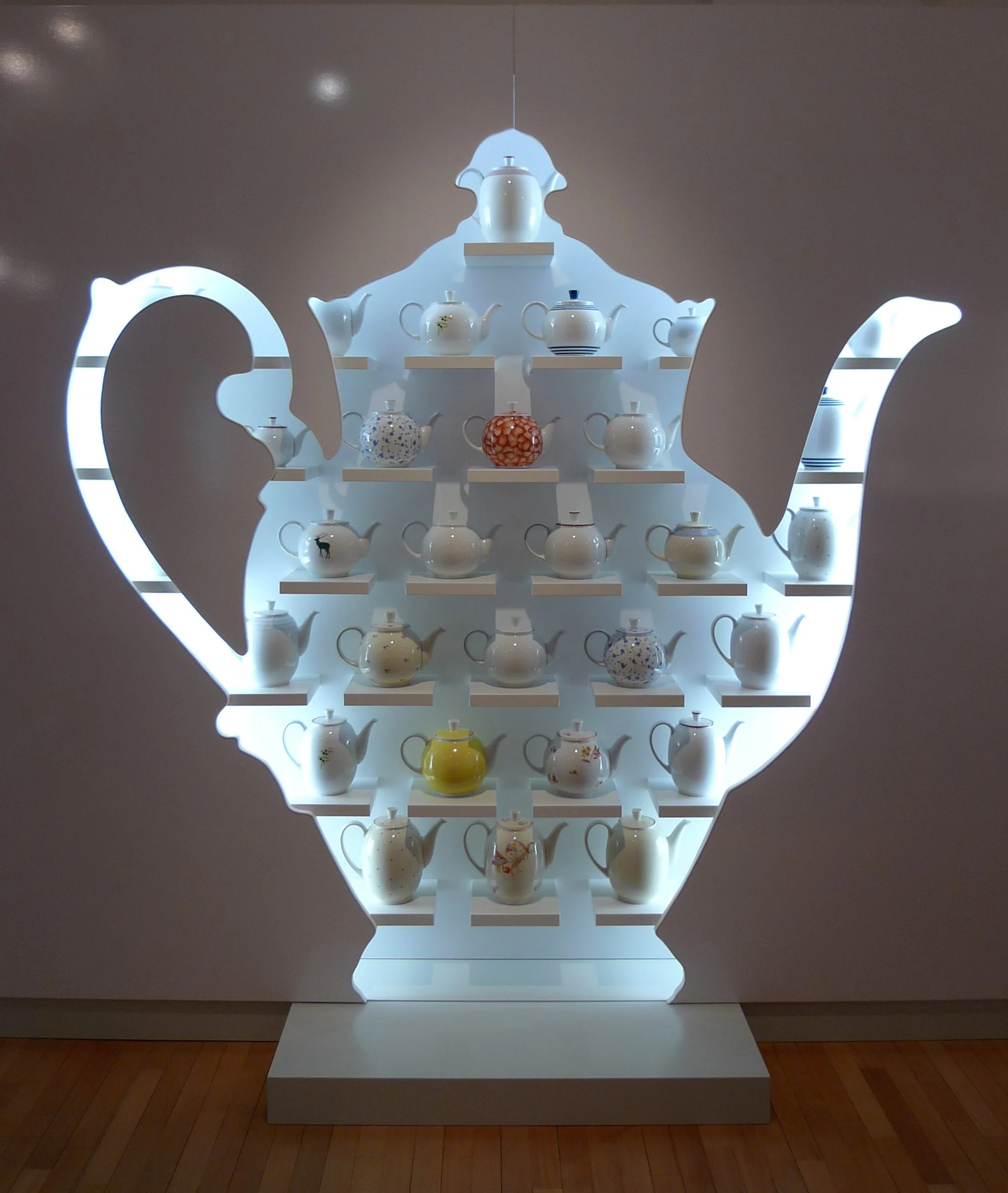 Ein leuchtendes Teekannen-Display im Ausstellungsraum, gestaltet in Form einer großen Teekanne mit inneren Regalen, die verschiedene Teekannen in unterschiedlichen Designs und Farben präsentieren.