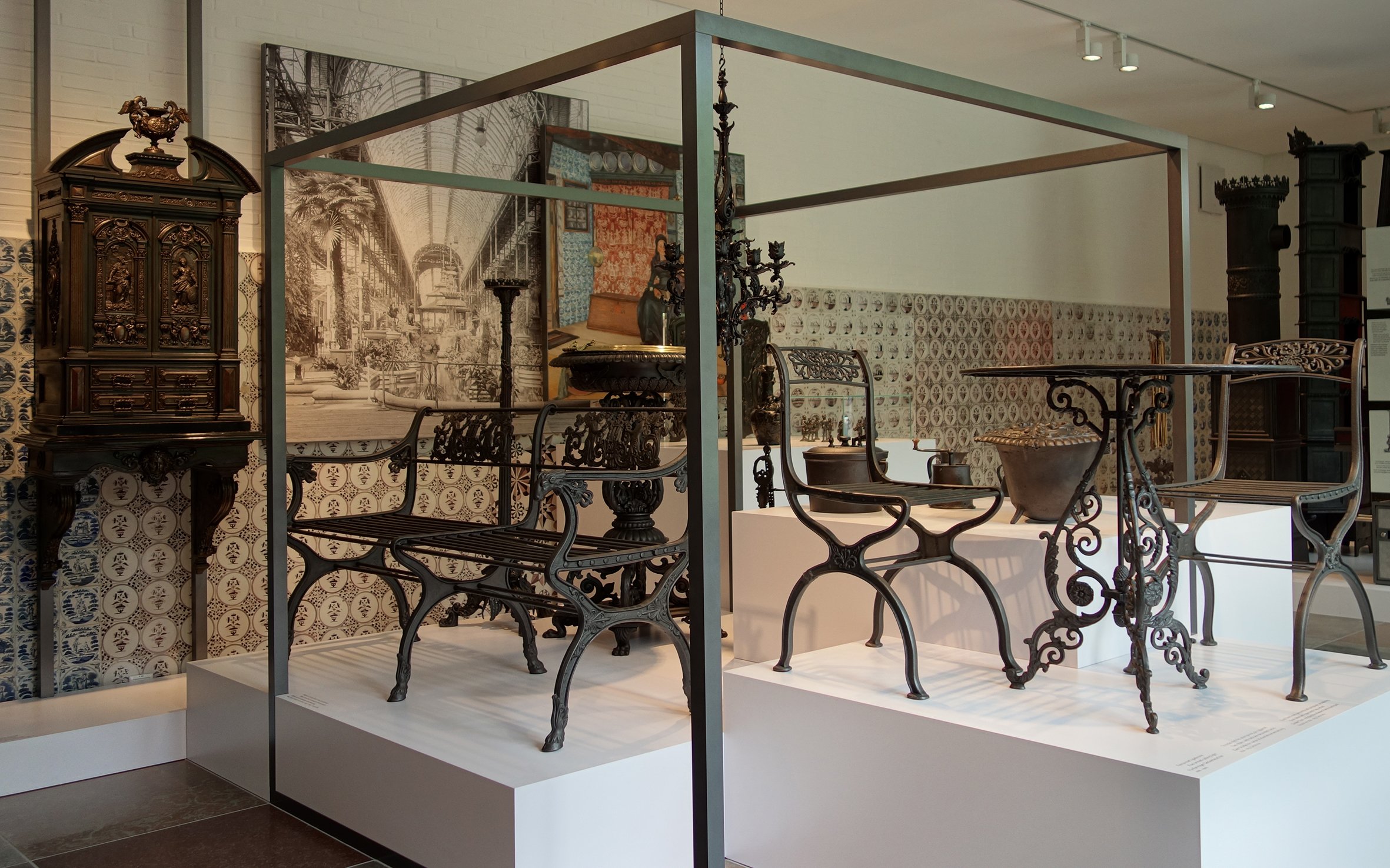 Verschiedene antike Metallmöbel und Dekorationsgegenstände in einem Museum, mit einer großen Schwarz-Weiß-Fotografie im Hintergrund.