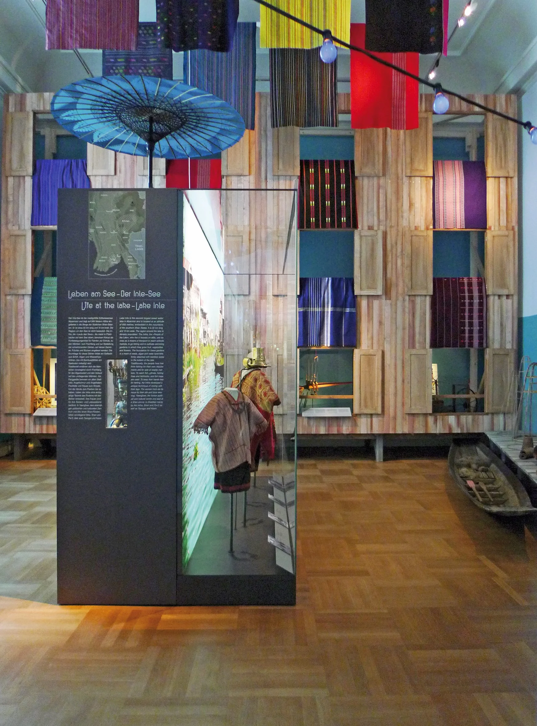 Ausstellung mit traditionellen Stoffen und Kleidungsstücken, Boote und Informationsstände, Dekoration mit bunten Stoffen und Lampen.