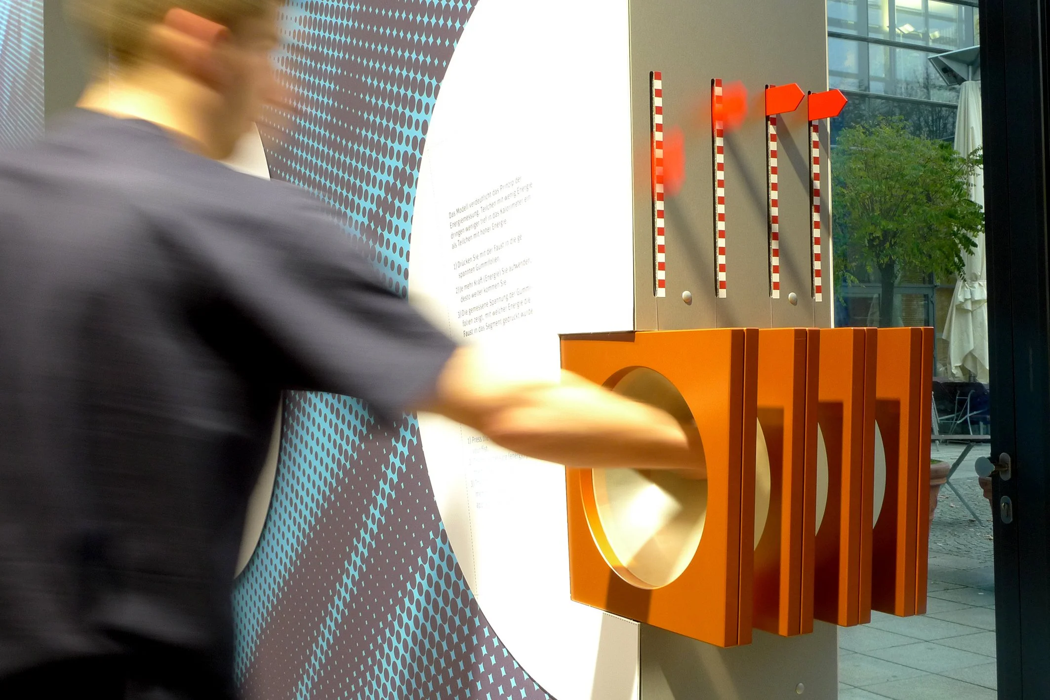 Eine Person drückt auf ein orangefarbenes Knöpfchen an einer interaktiven Ausstellung oder Übung, die vertikale Streifen und eine große orangefarbene Scheibe umfasst, im Hintergrund ist Glas mit Blick auf einen grünen Baum zu sehen.