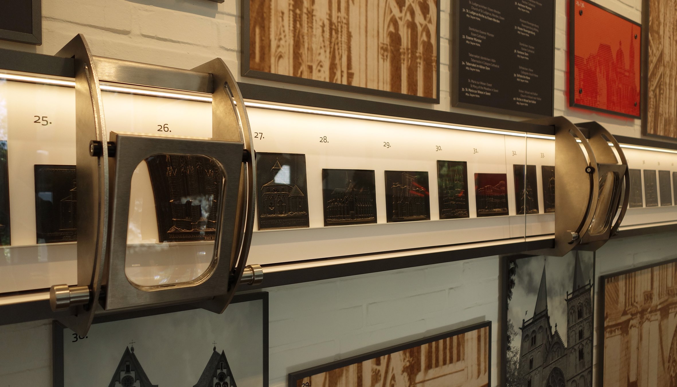 Ausstellungsdisplay mit nummerierten kleinen Modellen oder Reliefs in einem Museum, umgeben von Bildern und Texttafeln an der Wand.