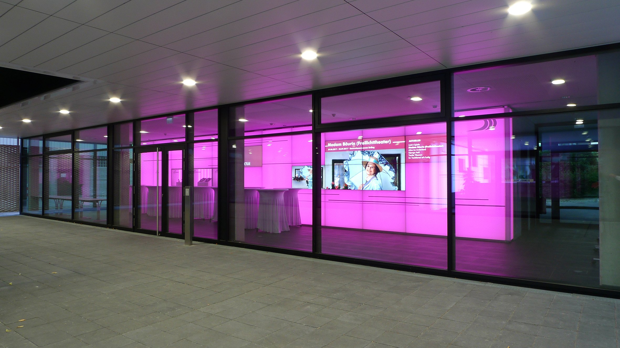Ein modernes Gebäude mit großen Glasfenstern, durch die ein pink beleuchteter Innenraum sichtbar ist. Im Inneren befinden sich digitale Bildschirme und eine Theke.