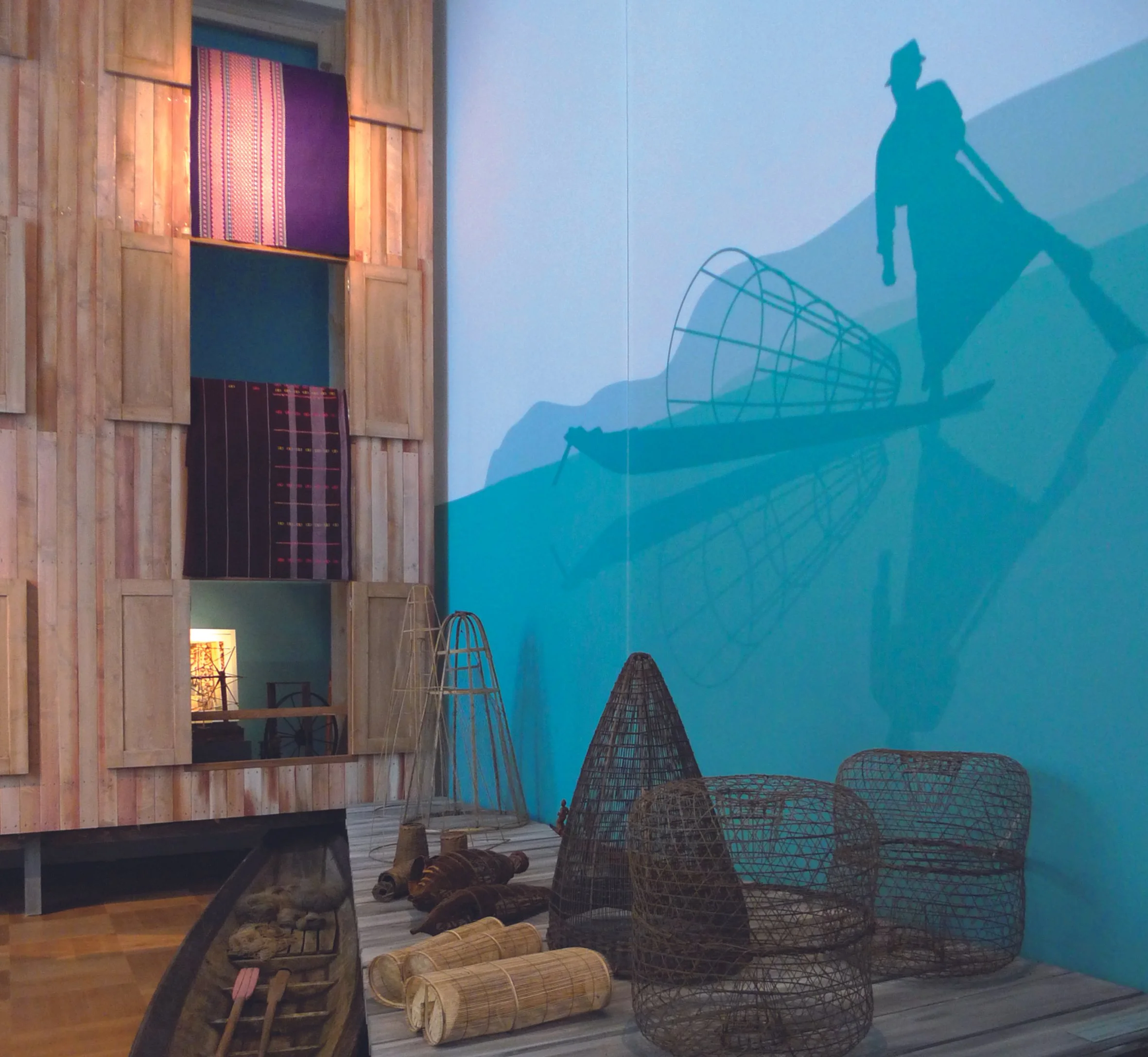 Eine Wandecke mit Holzpanelen, verschiedene Körbe und Holzgegenstände, sowie eine Wand mit blauer Farbe und einem Schatten eines Menschen, der auf einem Surfbrett steht.