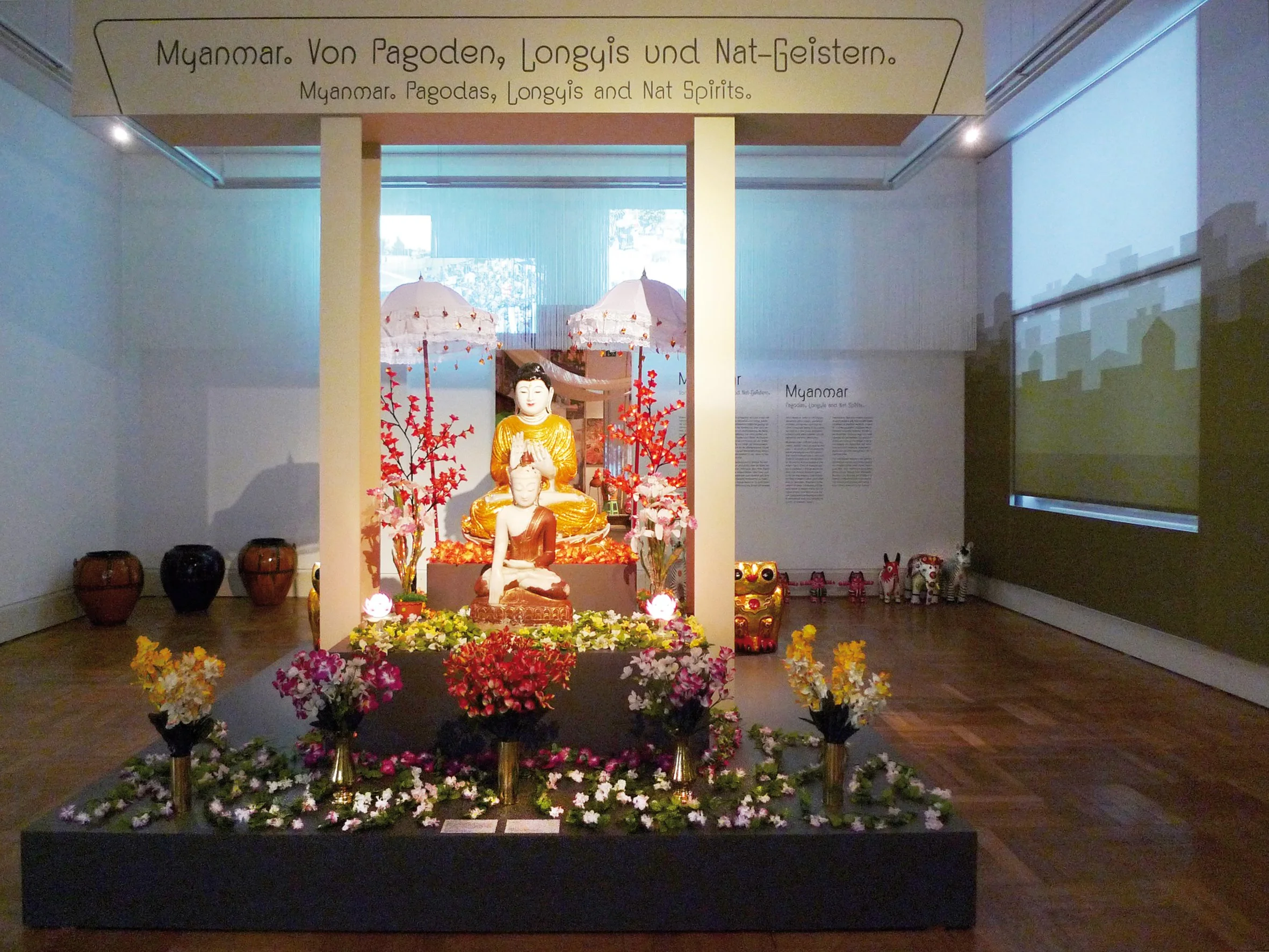 Buddhastatuen in einem Ausstellungssaal, dekoriert mit Blumen, roten Papierblumen und Teekannen. Ein Banner oben beschreibt Myanmar, Pagoden, Longys und Naturgeister auf Deutsch und Englisch.