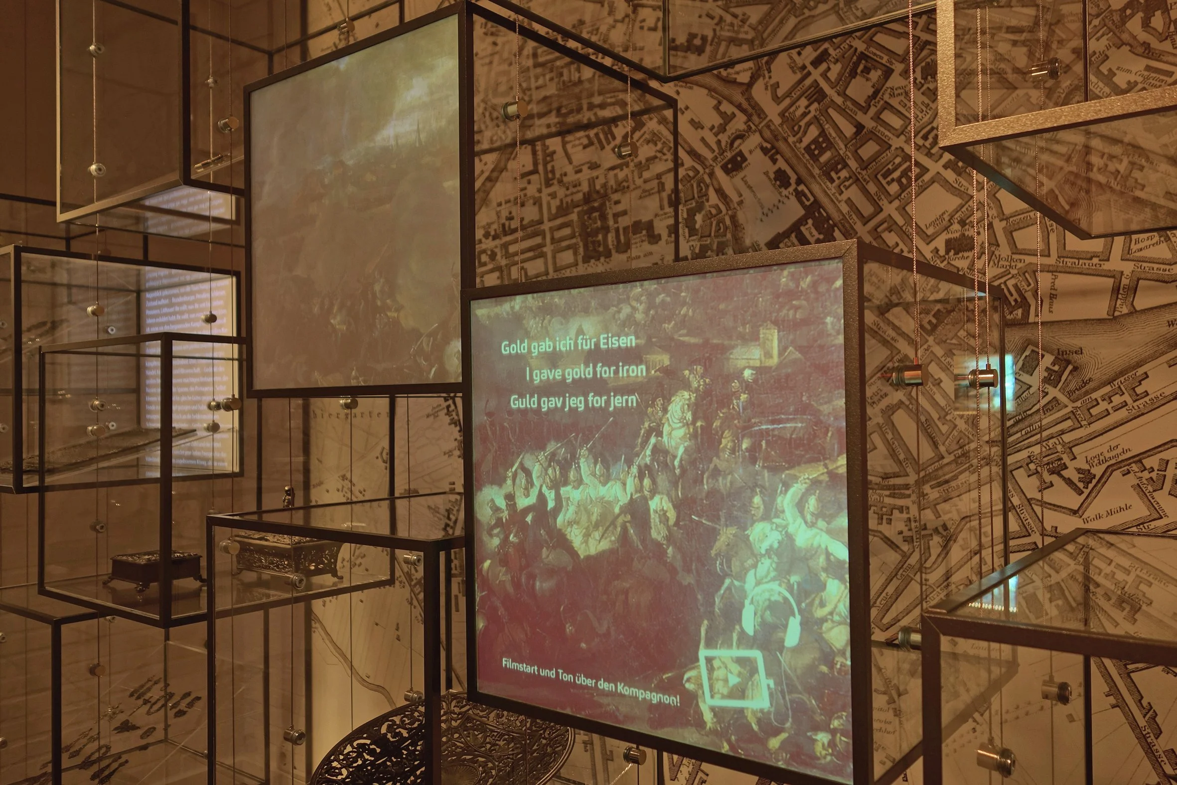 Ausstellung mit mehreren leeren, rahmenlosen Glasboxen, die an Drähten hängen, und einem Bildschirm mit historischen Bildern und deutschen Texten. Die Wand im Hintergrund ist mit einem Stadtplan gestaltet.