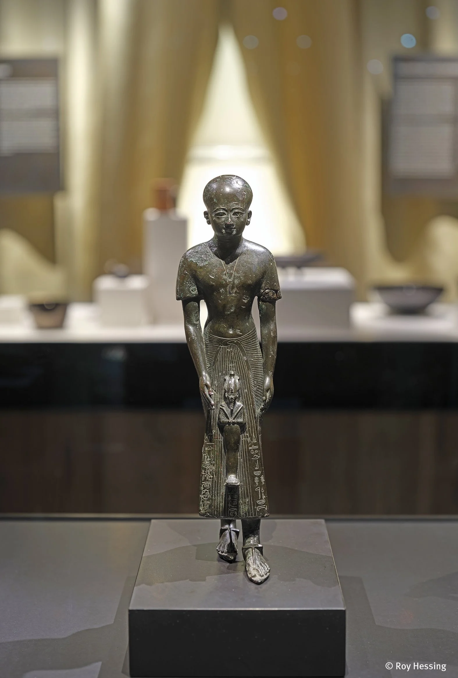 Ägyptische Statue eines jungen Mannes mit Halsschmuck, bekleidet mit einer Lendenschurft, stand auf einem schwarzen Sockel in einem Museum. Der Hintergrund ist unscharf mit goldfarbenen Vorhängen und Vitrinen.