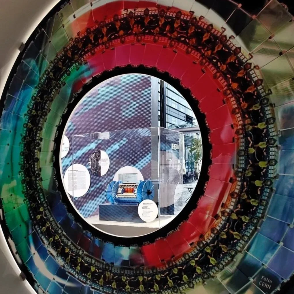 Ein detailreiches, farbenfrohes Kunstwerk, das aussieht wie eine große, runde Maschine oder Installation. Im Mittelpunkt steht ein transparentes Gehäuse mit einem mechanischen Gerät darin, möglicherweise ein Kunst- oder Technikobjekt. Das Kunstwerk ist von farbigen, kreisförmigen Schichten aus Plastik oder Glas umgeben, die in Regenbogenfarben angeordnet sind. Im Hintergrund sind Infos und digitale Displays, vermutlich in einem Museum oder Ausstellungsraum.