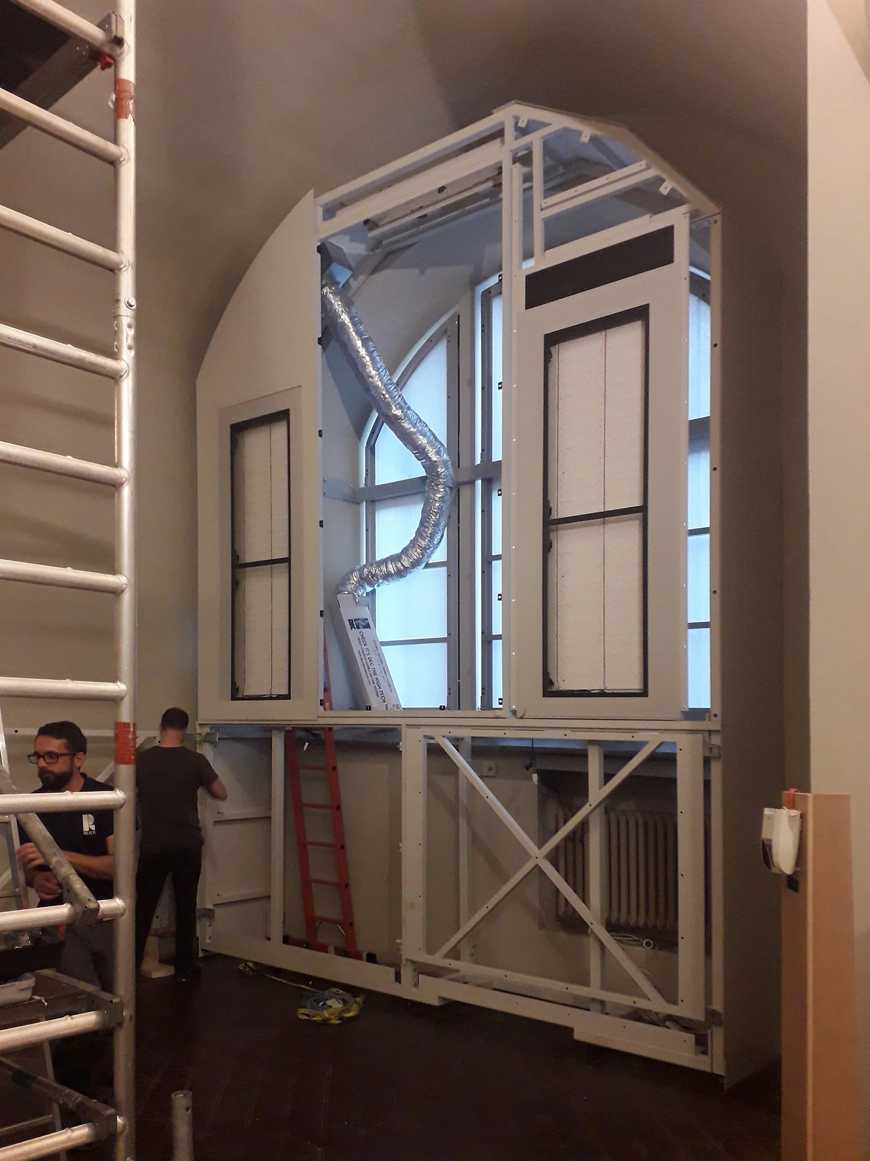 Ein Baugerüst vor einer großen Fensteröffnung, die gerade im Ausbau ist. Es sind Menschen bei der Arbeit, ein roter Leiter steht im Raum, und eine flexible Luftleitung ist sichtbar.