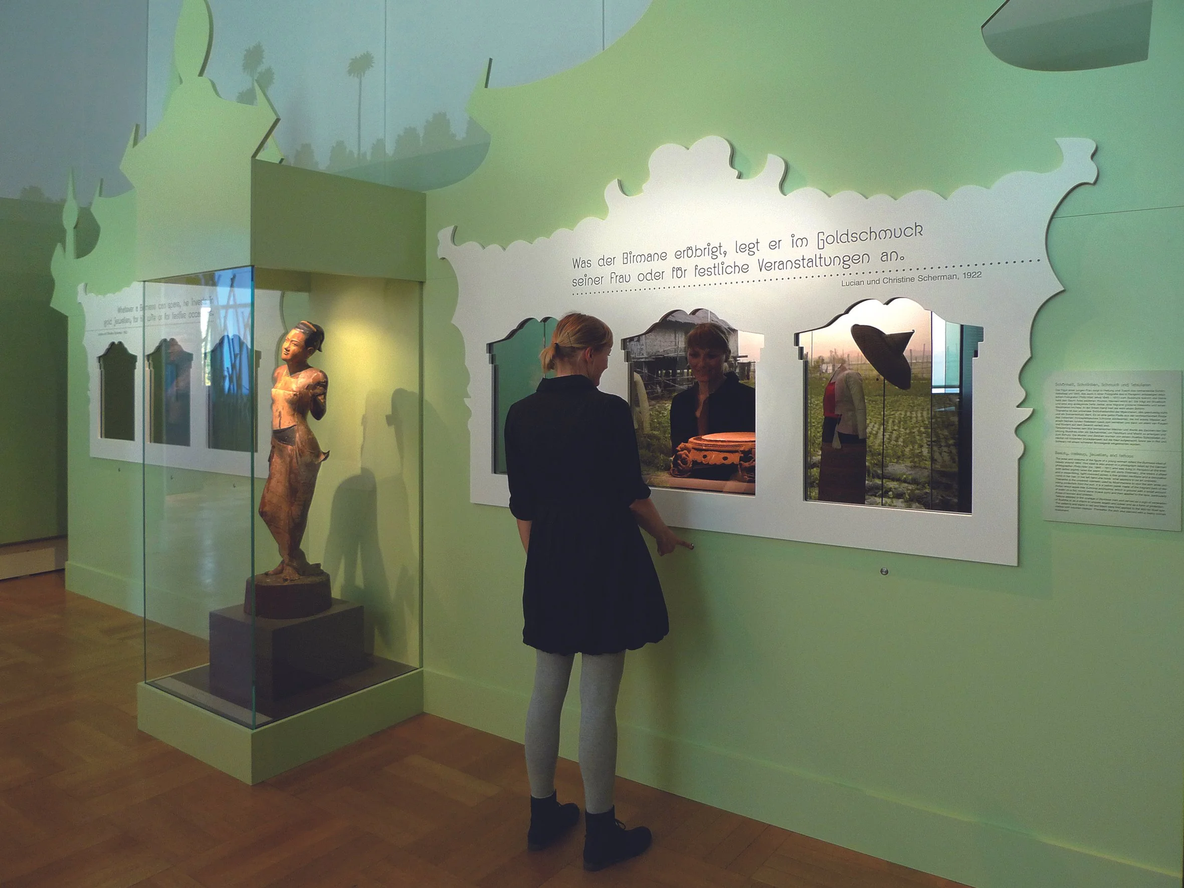 Zwei Frauen betrachten eine Ausstellung in einem Museum. Es gibt eine Statue und eine Wand mit Ausschnitten und Texten.