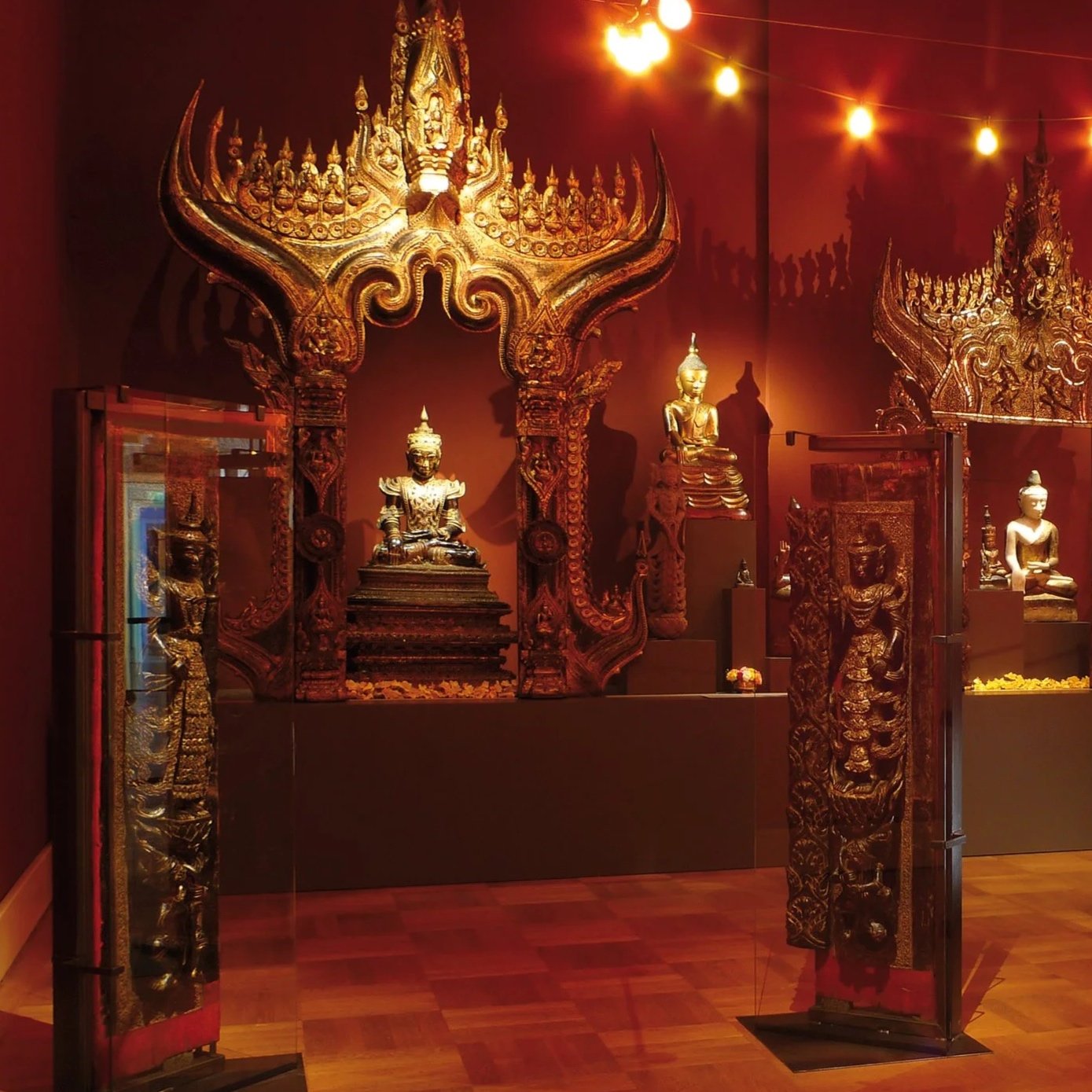 Goldene Buddha-Statuen und traditionelle thailändische Kunstwerke in einem Museumsraum mit warmem Licht.