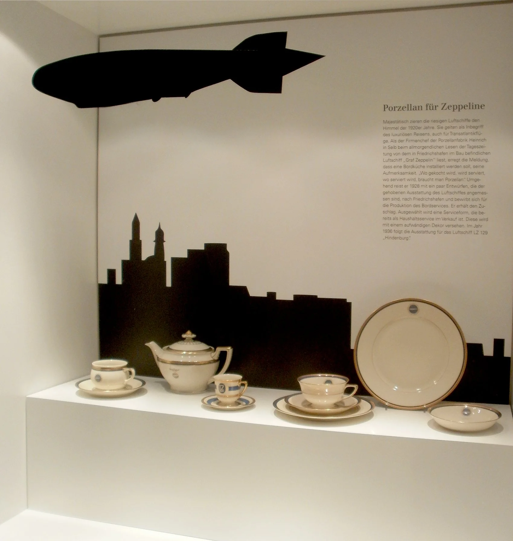 Porzellanservice mit Kaffee- und Teetassen, Untertassen, Zucker- und Milchkännchen auf einer weißen Ausstellungsfläche. Hintergrund mit Silhouette einer Stadt und einem schwarzen Zeppelin. Beschreibung zum Porzellan für Zeppelin im Deutschen.