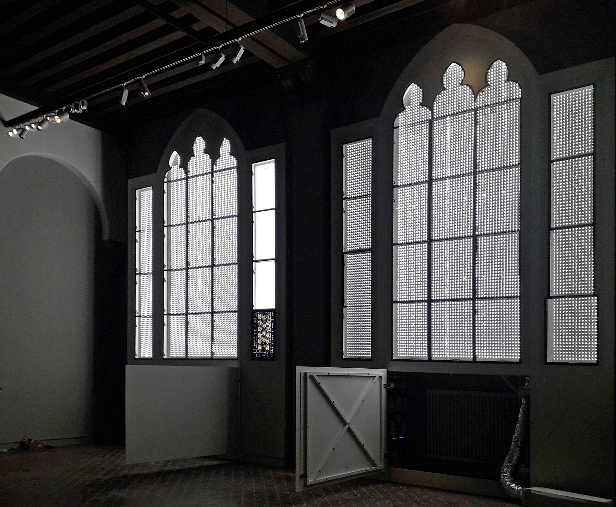 Innenansicht eines Raumes mit großen gotischen Fenster, die mit perforiertem Metall bedeckt sind, und einer teilweise geöffneten weißen Tür, auf dem Boden liegt ein Teppich.