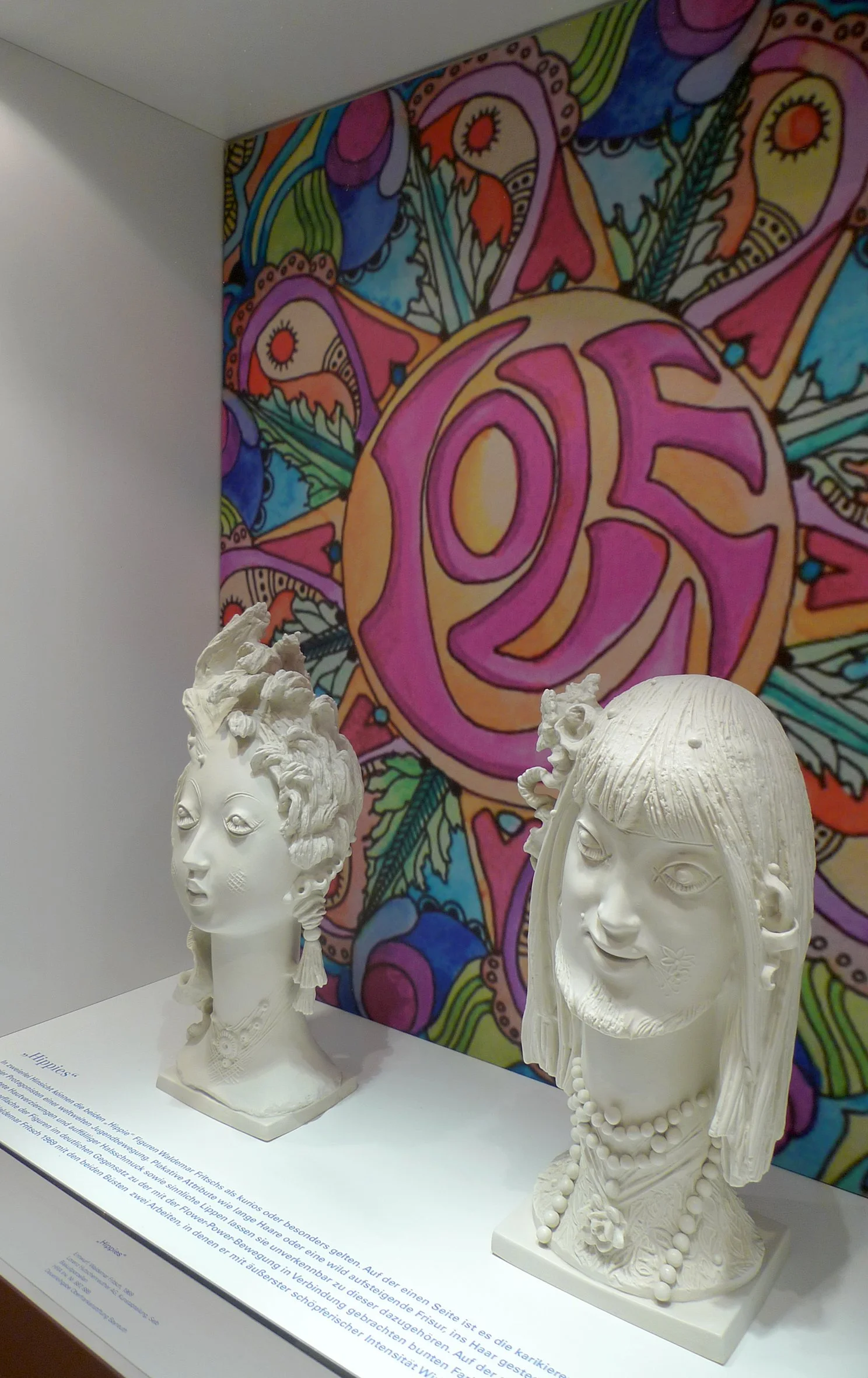 Zwei weiße Skulpturen in einer Vitrine vor einem bunten, abstrakten Wandbild mit dem Wort "LOVE" in der Mitte.