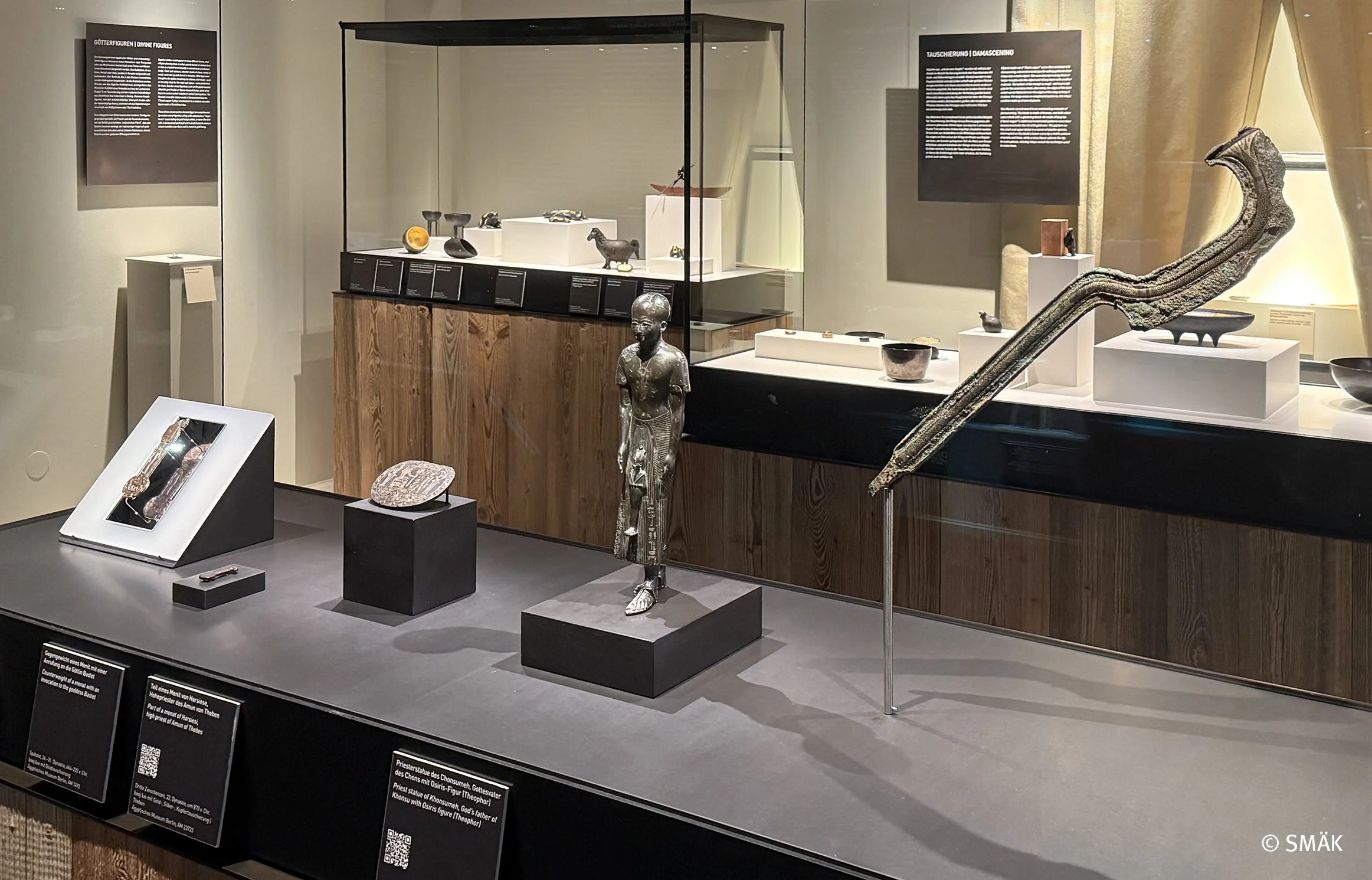 Ausstellung in einem Museum mit verschiedenen antiken Artefakten, darunter eine Bronzestatuette einer menschlichen Figur, eine größere geschnitzte Figur, eine lange, gebogene Beinknochen mit einer drehbaren Unterstützung, verschiedene kleinere Gegens