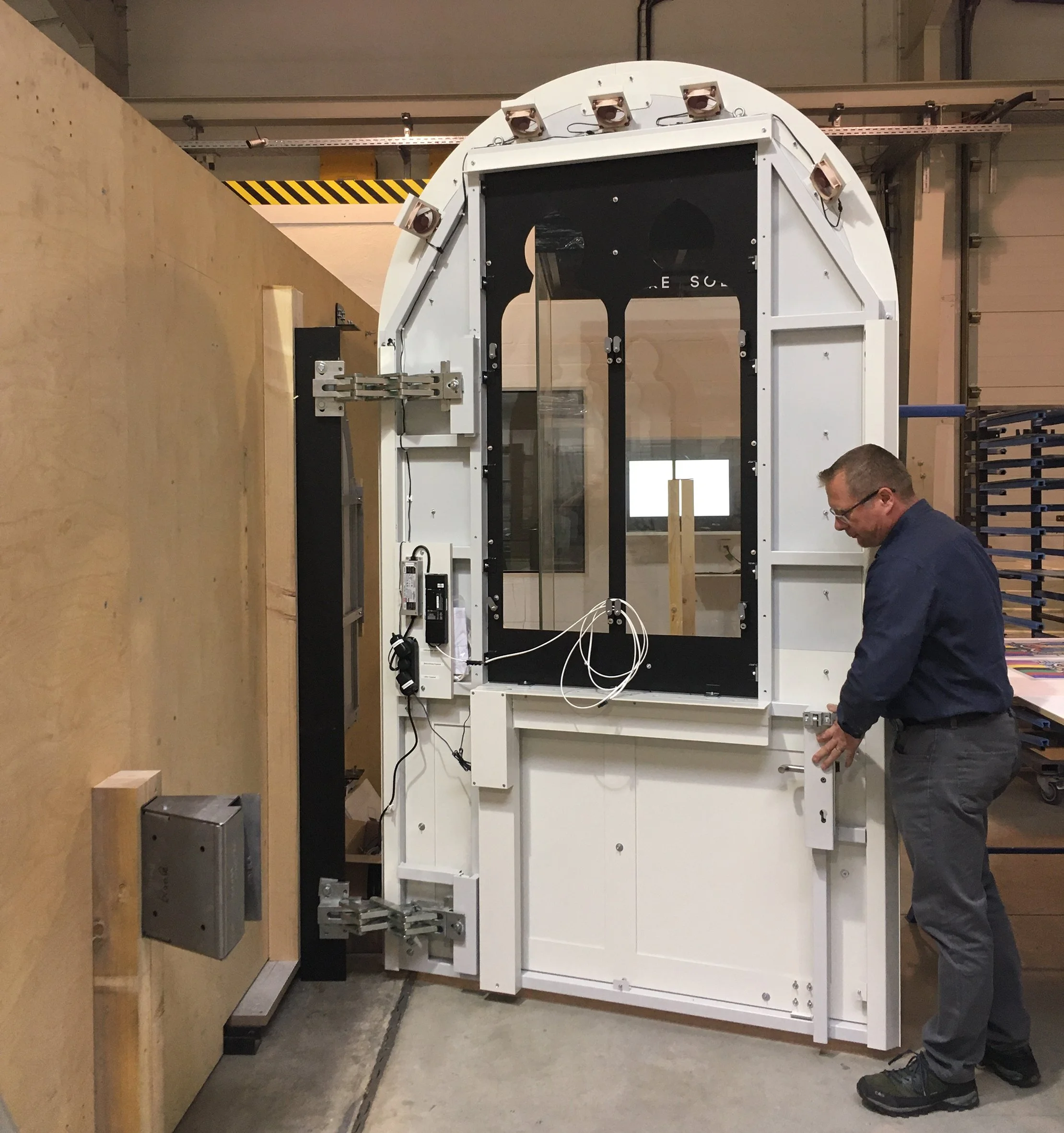 Mann arbeitet an einer großen, isolierten Kabine, vermutlich für Tests oder Forschung, in einer Werkstatt oder Fabrikhalle.