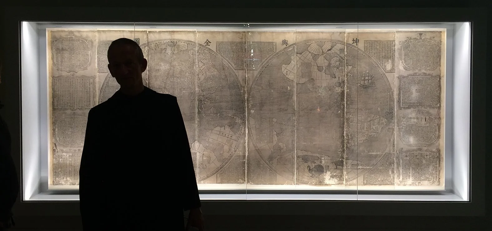 Begleitete Person vor antikem Weltkarten- oder Landkarten-Relief im Museum, in einem beleuchteten Rahmen vor dunklem Hintergrund.