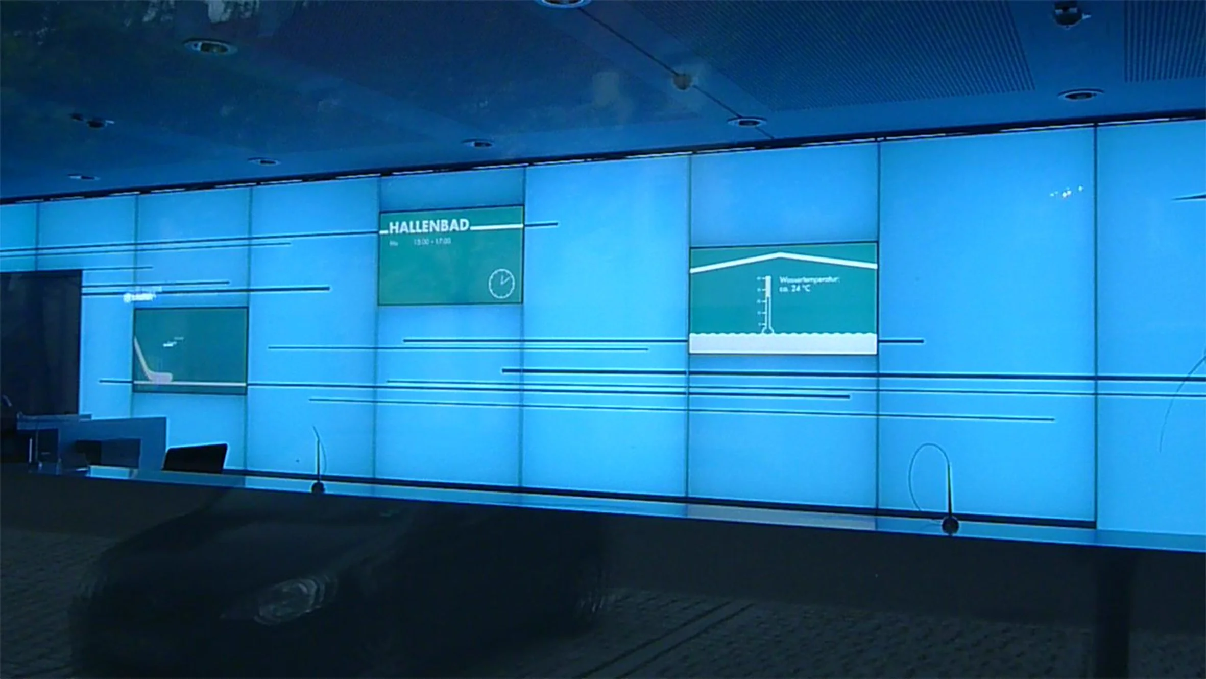 Eine große digitale Anzeige in einem Eingangsbereich zeigt Informationen über eine Hallenbadöffnung und die Temperatur an.