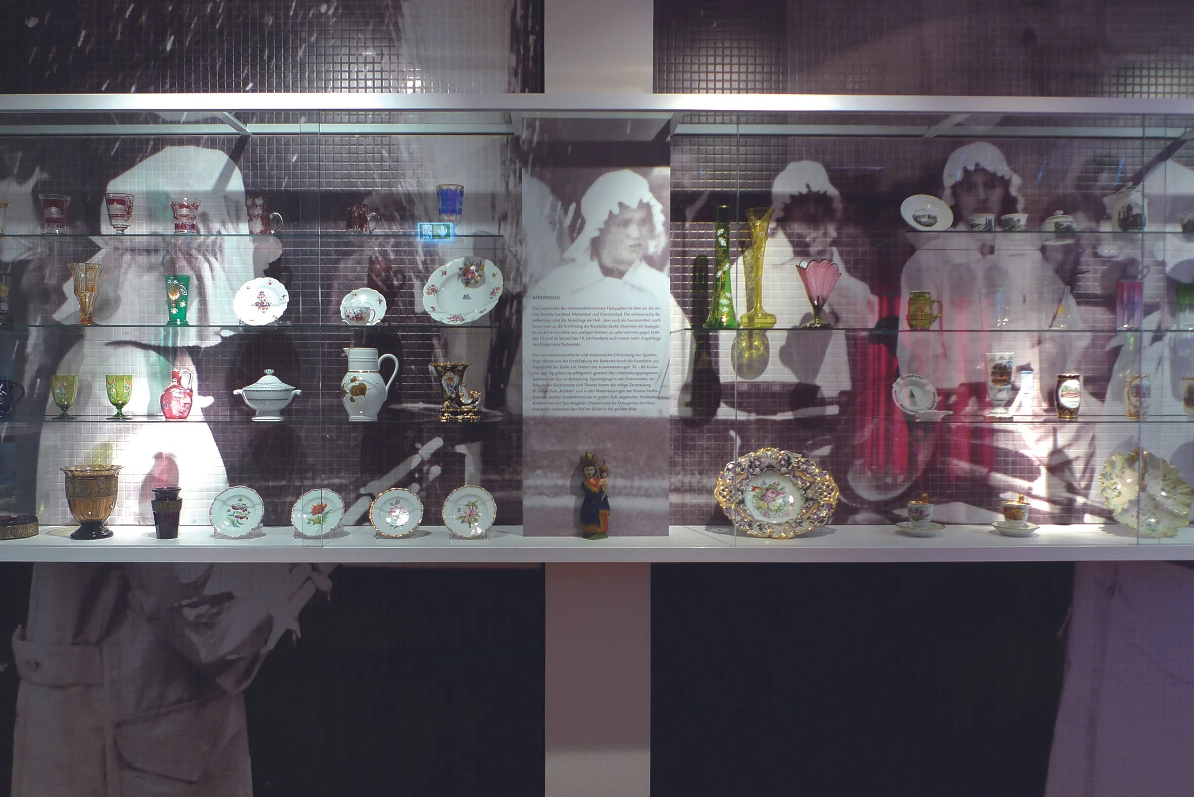 Ansicht einer Vitrine mit Porzellan, neben einem Radfahrermotiv mit Text. Im Hintergrund zwei Frauen in Trachten, die Porzellan sammeln.