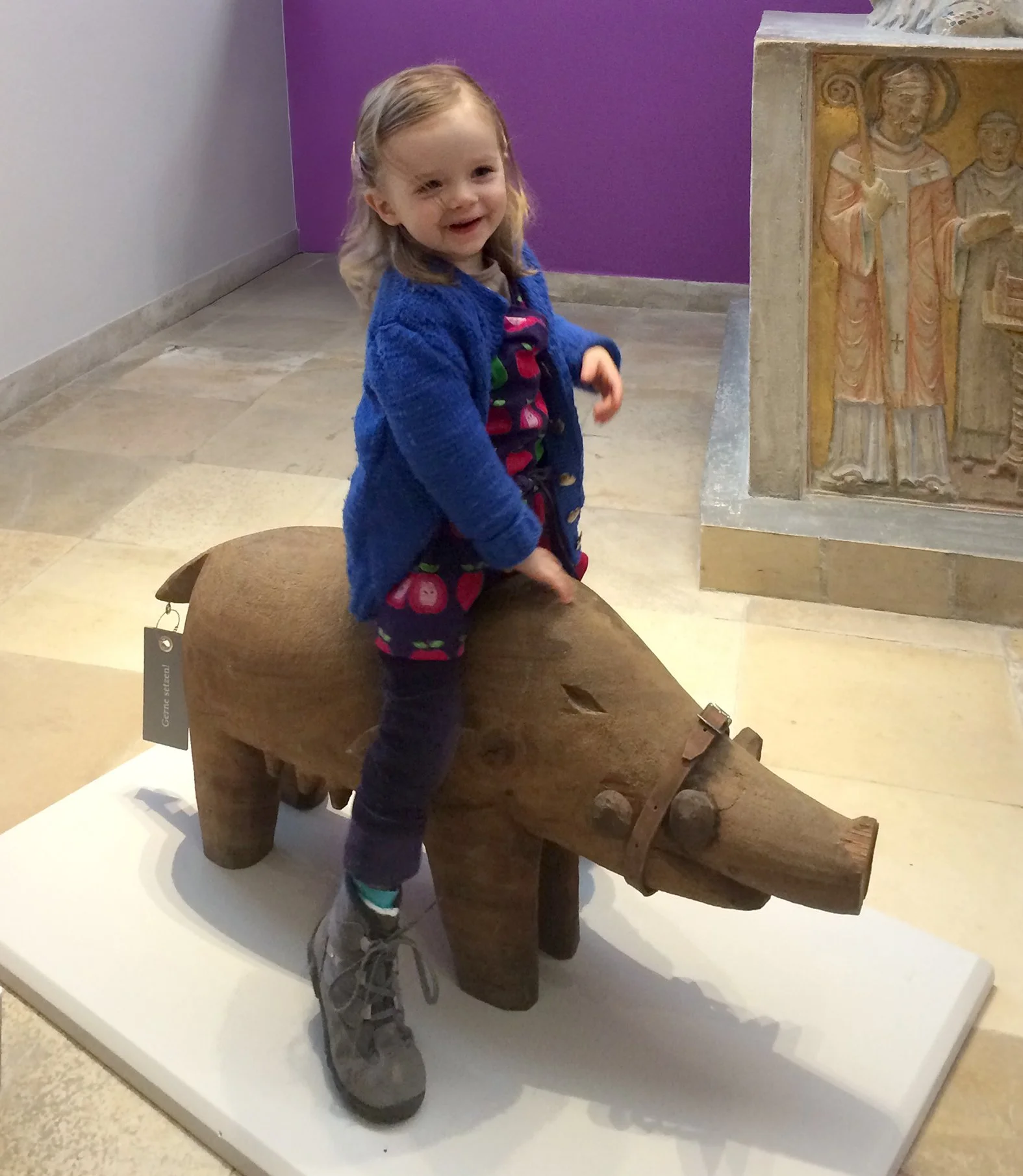 Ein kleines Mädchen sitzt auf einer Holzfigur eines Wildschweins, in einem Museum oder Ausstellung mit Kunstwerken im Hintergrund.