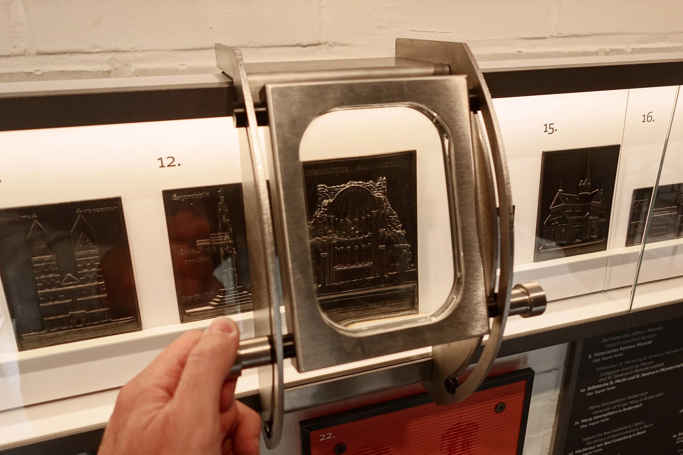 Person hält eine Lupe vor eine silberne Plakette mit architektonischer Darstellung, im Hintergrund sind weitere Plaketten mit Gebäuden, die in einer Vitrine ausgestellt sind.