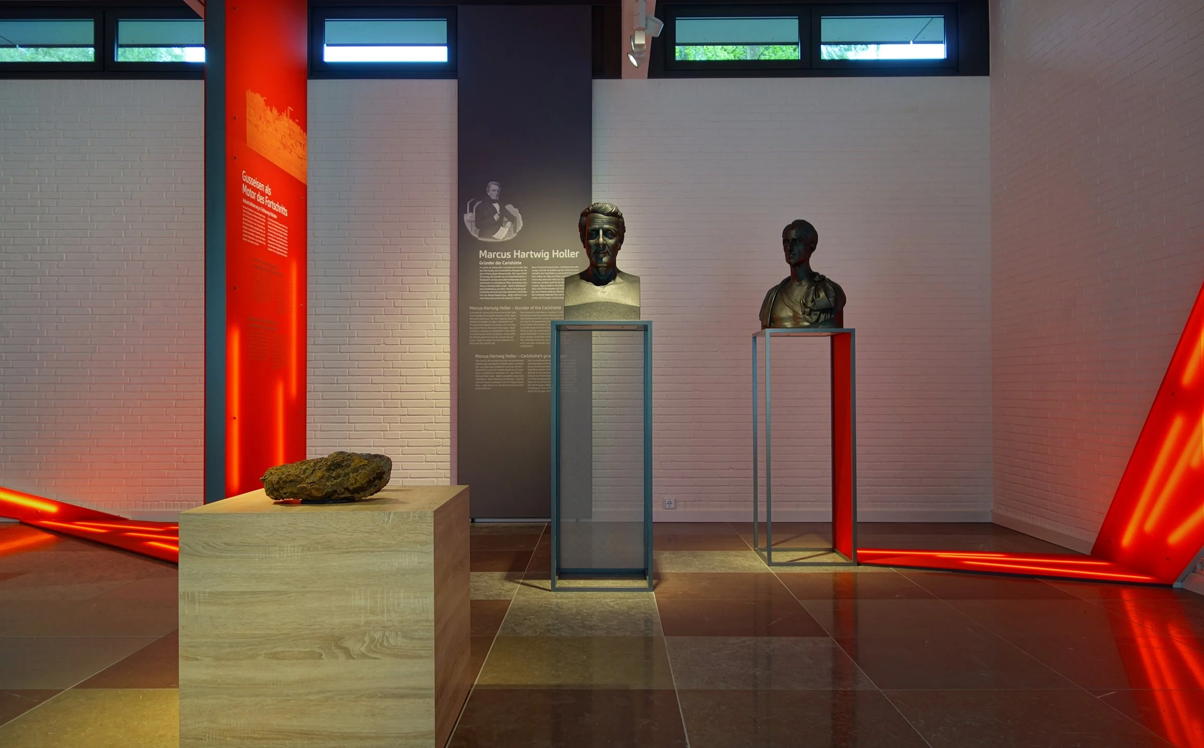 Ausstellung im Museum mit zwei Bronzestatuen, einem Felsen auf einer Holzplattform, einem schwarzen Info-Tafel mit Text, rotem LED-Licht und weißen Wänden.