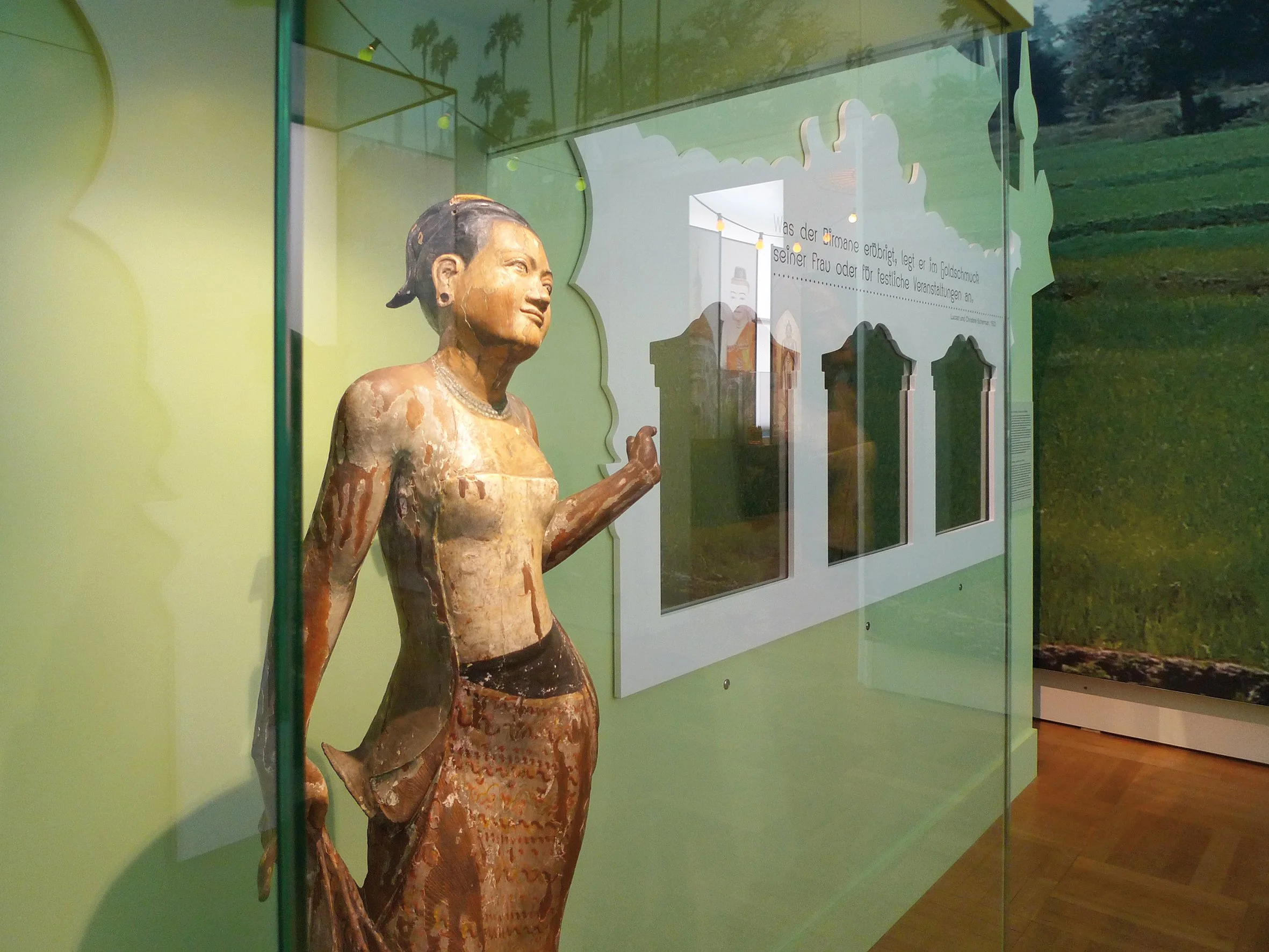 Eine alte, bemalte Holzfigur eines asiatischen Frauenbildes in einer Vitrine, mit Hintergrund einer grünen Wand und einer Wandazeige auf der rechten Seite.