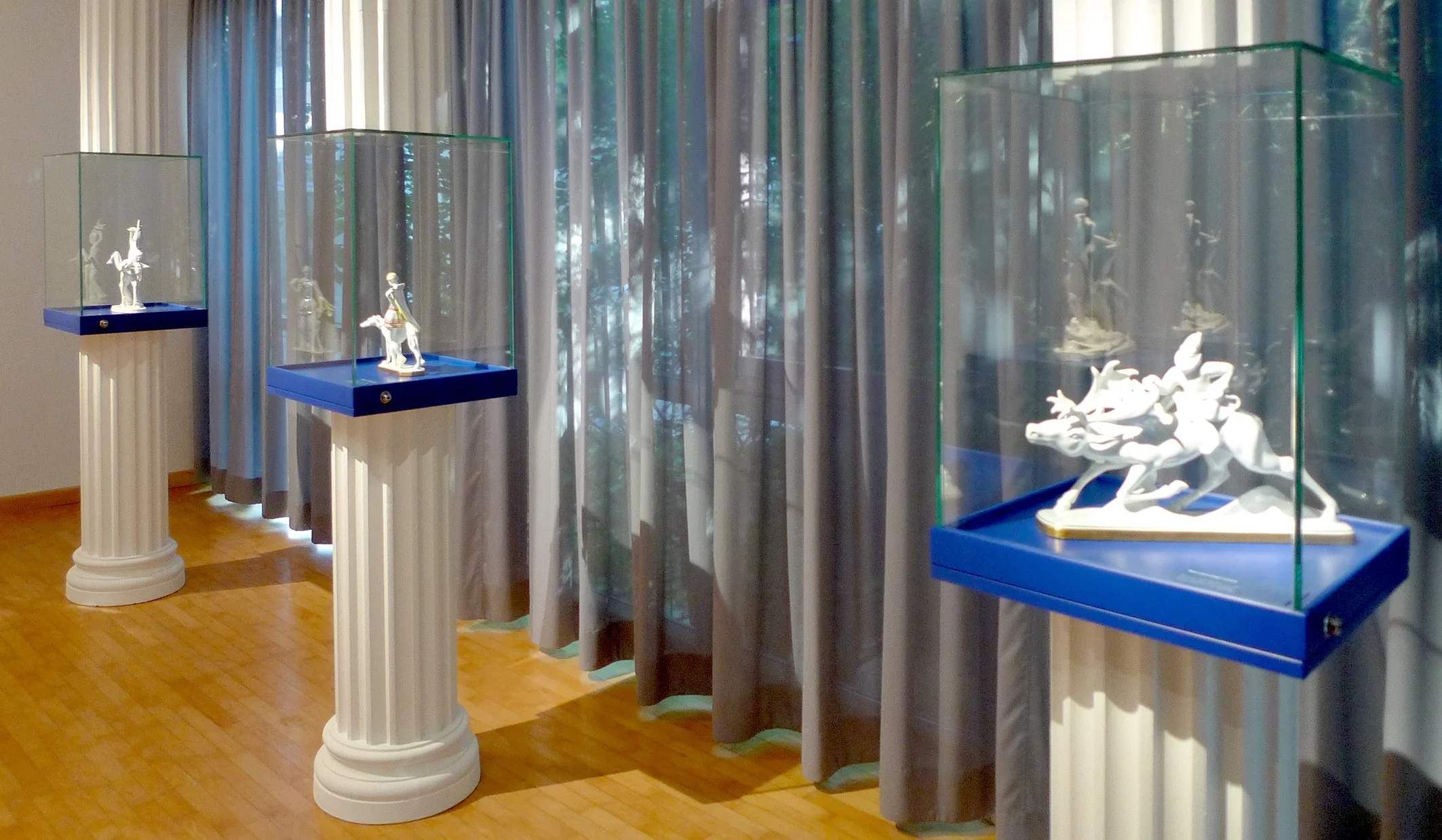 Drei Skulpturen in Glasvitrinen auf weißen Säulen im Museum, mit Vorhängen im Hintergrund.