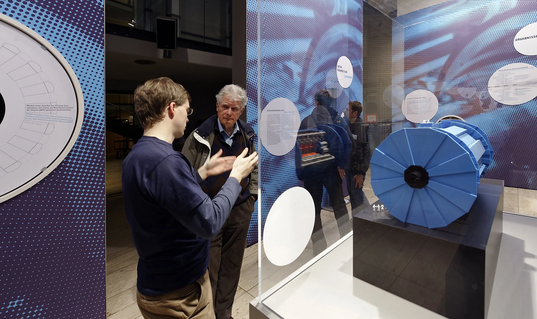 Zwei Männer sprechen vor einer Vitrine mit einem blauen mechanischen Gerät, vermutlich ein wissenschaftliches Exponat, in einer Ausstellung.
