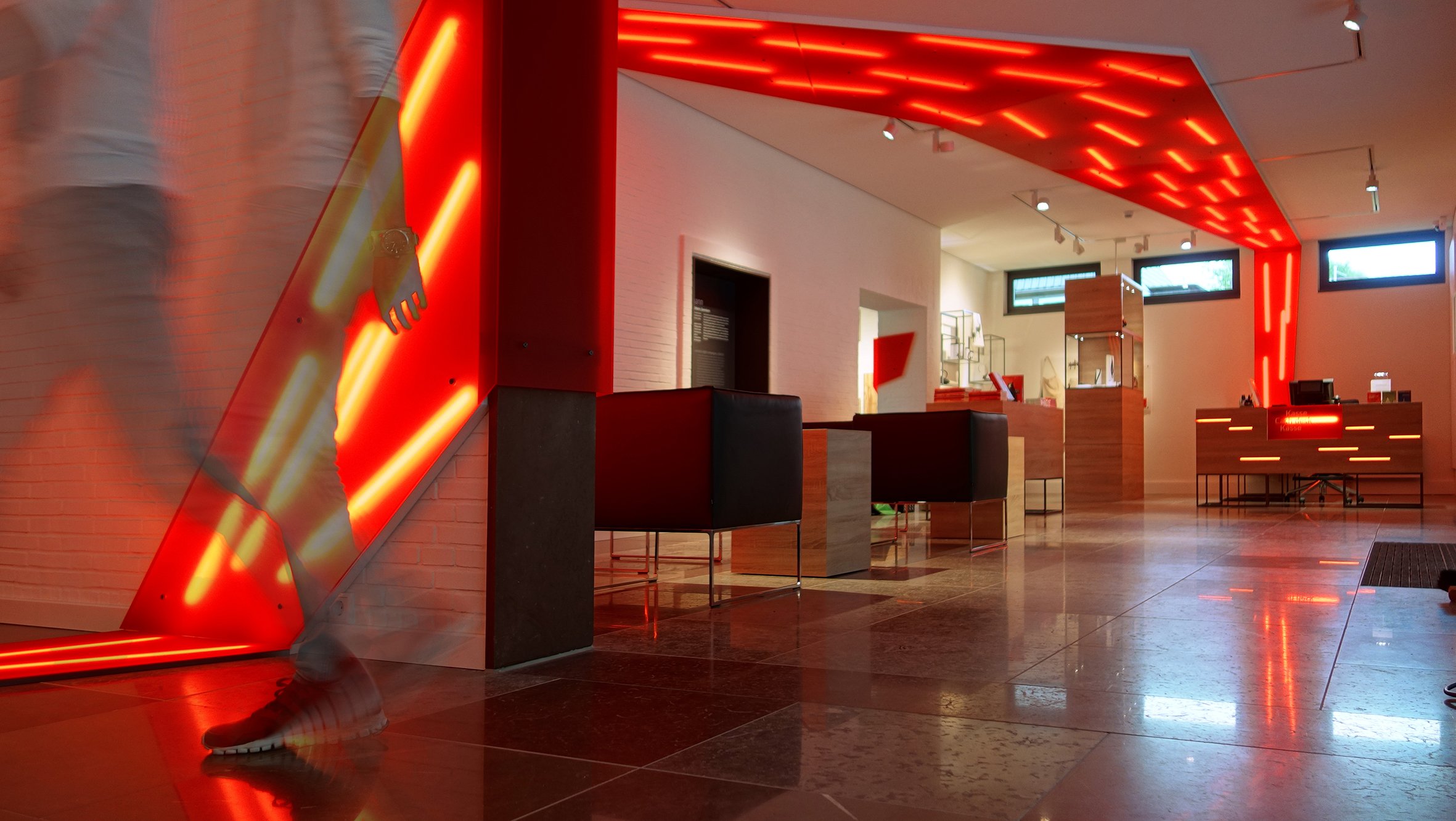 Innenaufnahme eines modernen Gebäudes mit roter Beleuchtung, Empfangstresen und Wartebereich, mehrere Fenster an den Wänden.