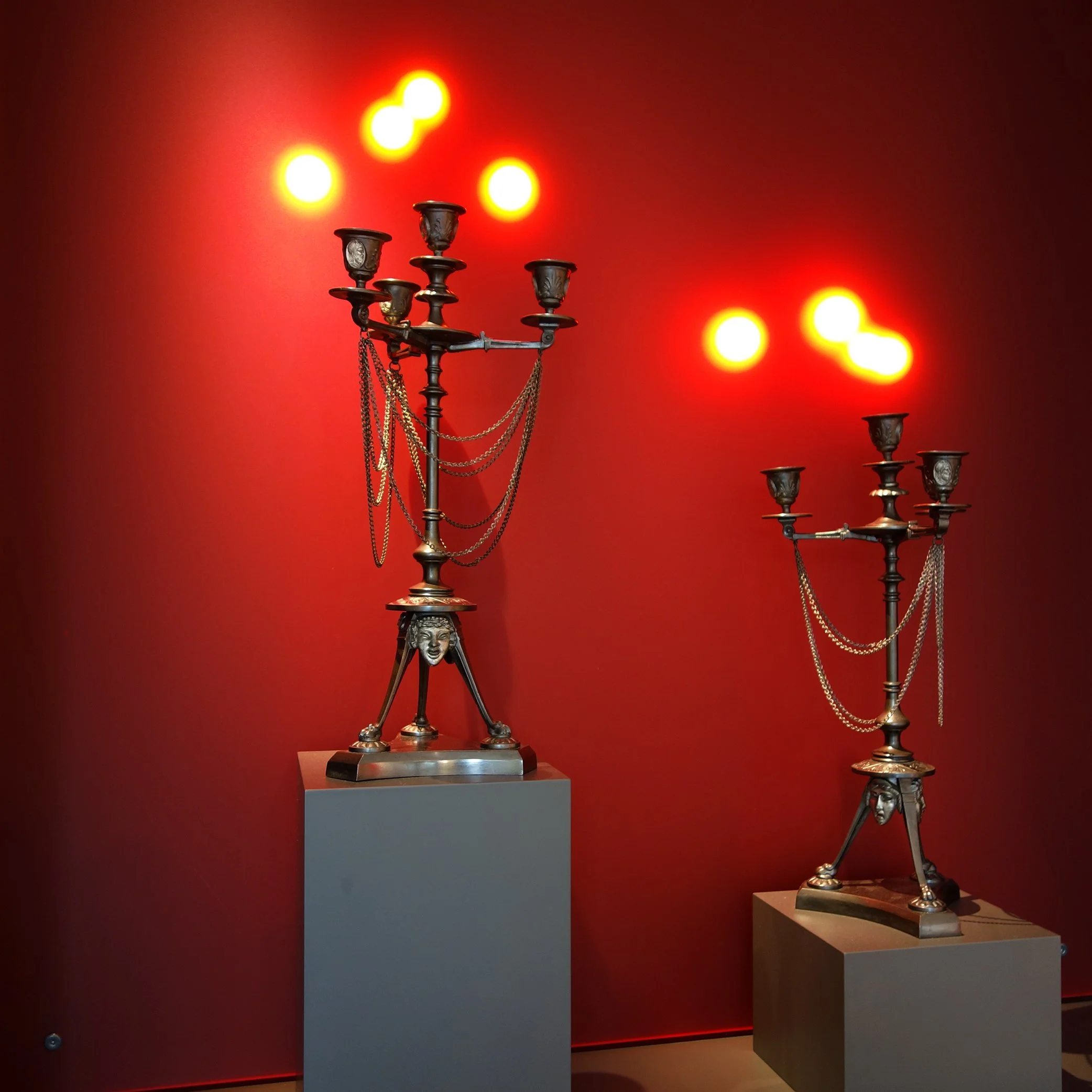 Zwei antike Laternenleuchter aus Metall auf Museumsplattformen, beleuchtet mit warmem Licht im Hintergrund.