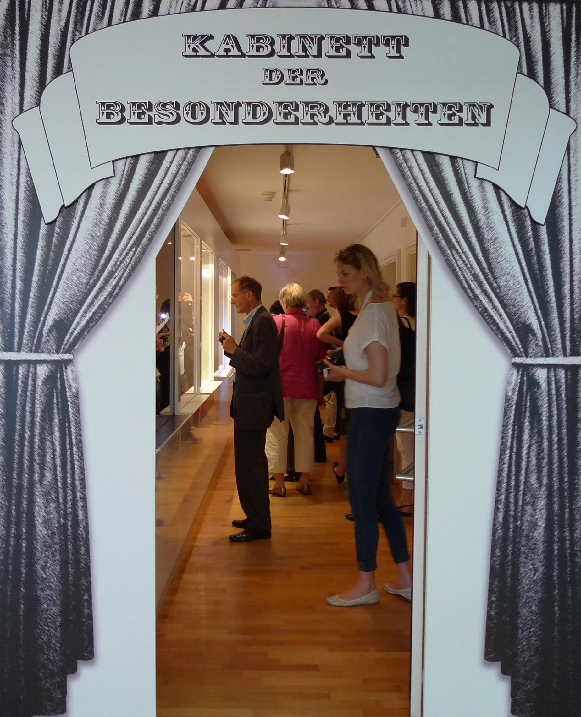 Menschen betrachten Ausstellungsstücke durch Schaufenster in einer Ausstellung, die Eingangstür ist mit einem Bühnenvorhang und einem Schild "Kabinett der Besonderheiten" dekoriert.