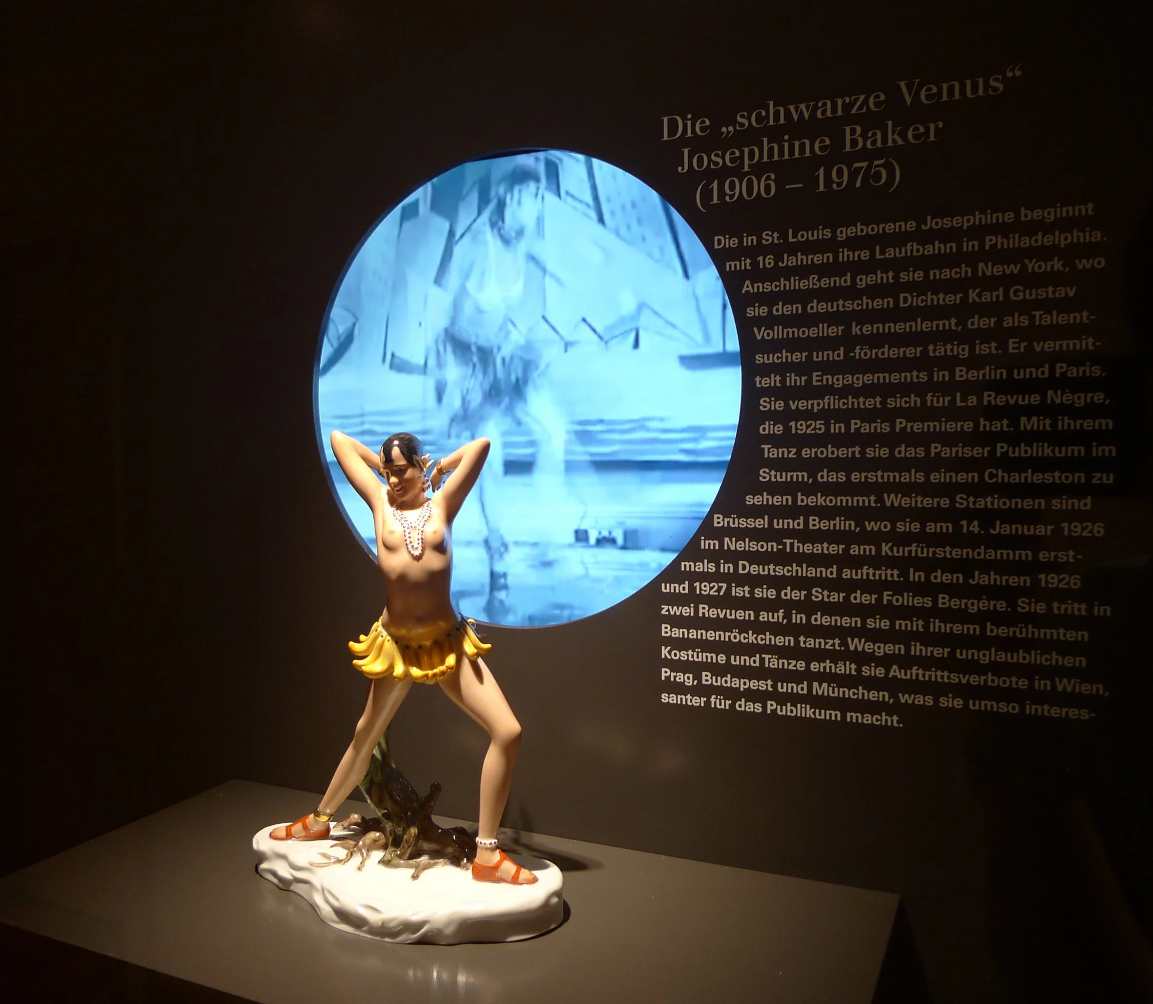 Eine Skulptur einer Tänzerin mit Banane-Kostüm, die auf einer weißen Plattform steht, im Hintergrund ein blauer Kreis mit einem schwarzen Hintergrund, daneben deutscher Text über die Künstlerin Josephine Baker.