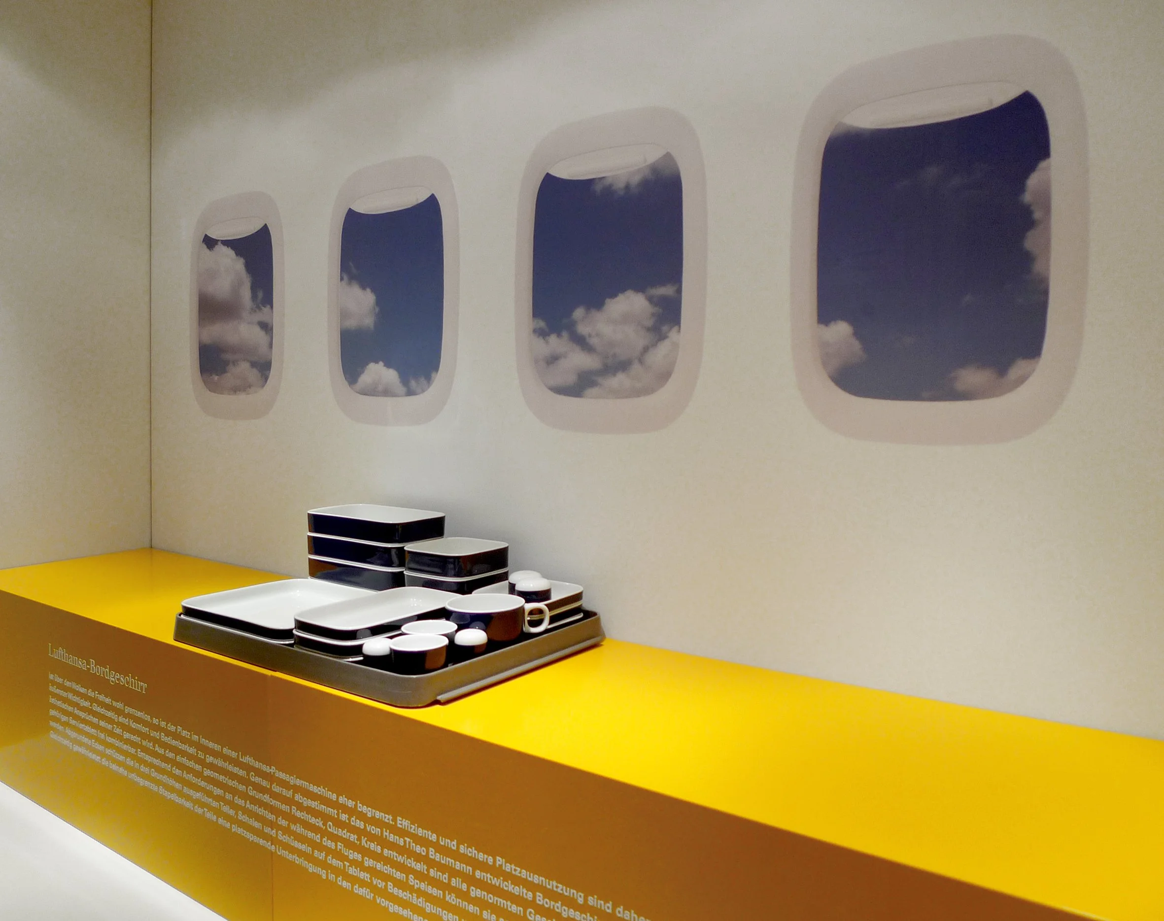 Ausstellung mit Maßen für einen Flugzeugtreppengeländer, mit Schwarz-Weiß-Geschirr auf gelbem Tisch und Fenstern, die den Himmel mit Wolken zeigen.