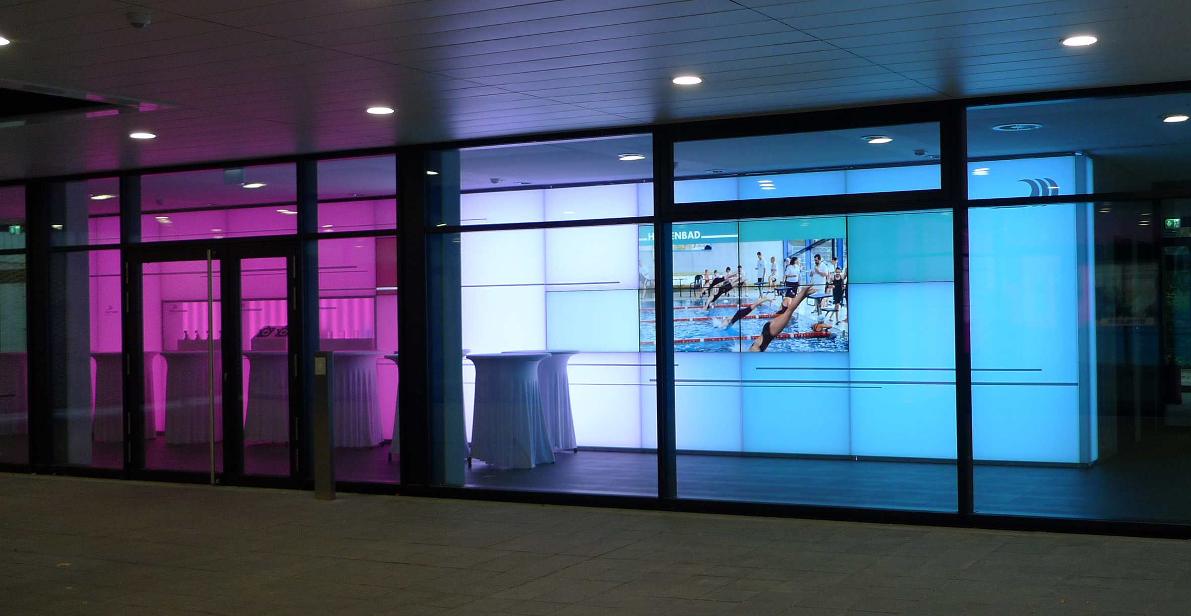 Fassadenspiegel mit buntem LED-Licht und einem großen Digitalbildschirm, der eine Schwimmbad- oder Wettkampfaufnahme zeigt.