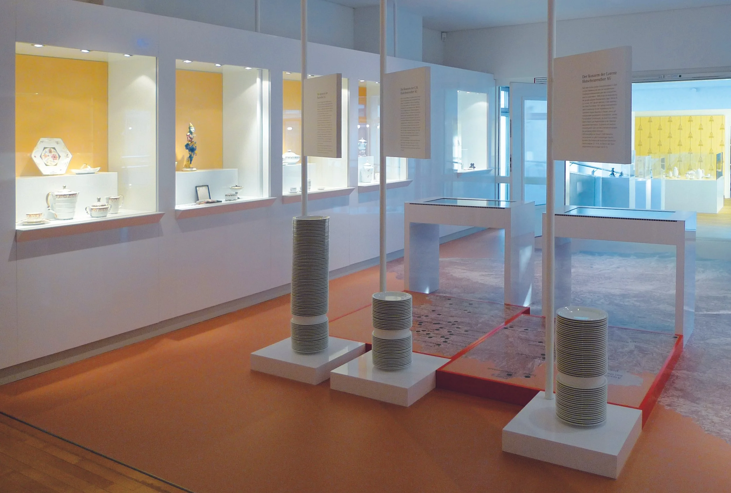 Ausstellung im Museum mit Porzellan und Texttafeln, weiße Vitrinen und Standfüße, orangefarbener Boden, helle Wände, große Fenster