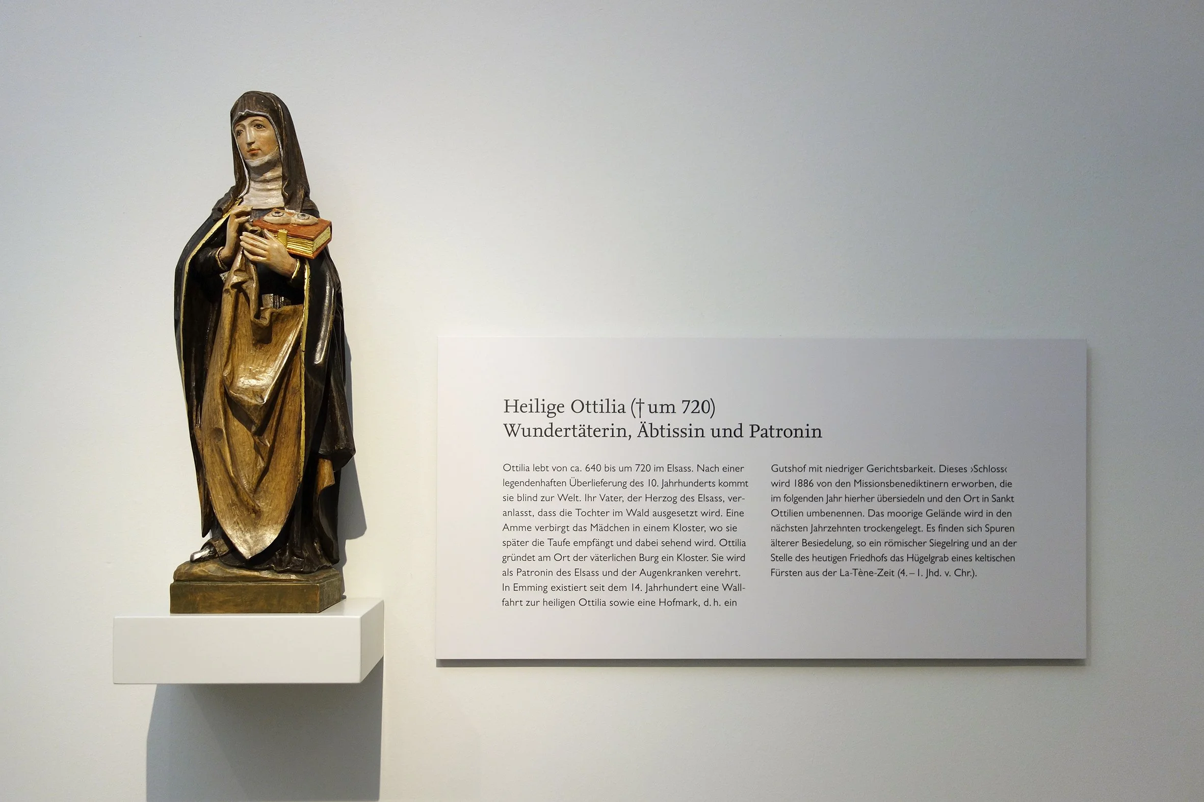 Skulptur der Hl. Ottilia, die ein Buch und eine Kette hält, mit einem Erklärungsschild auf Deutsch neben der Figur.