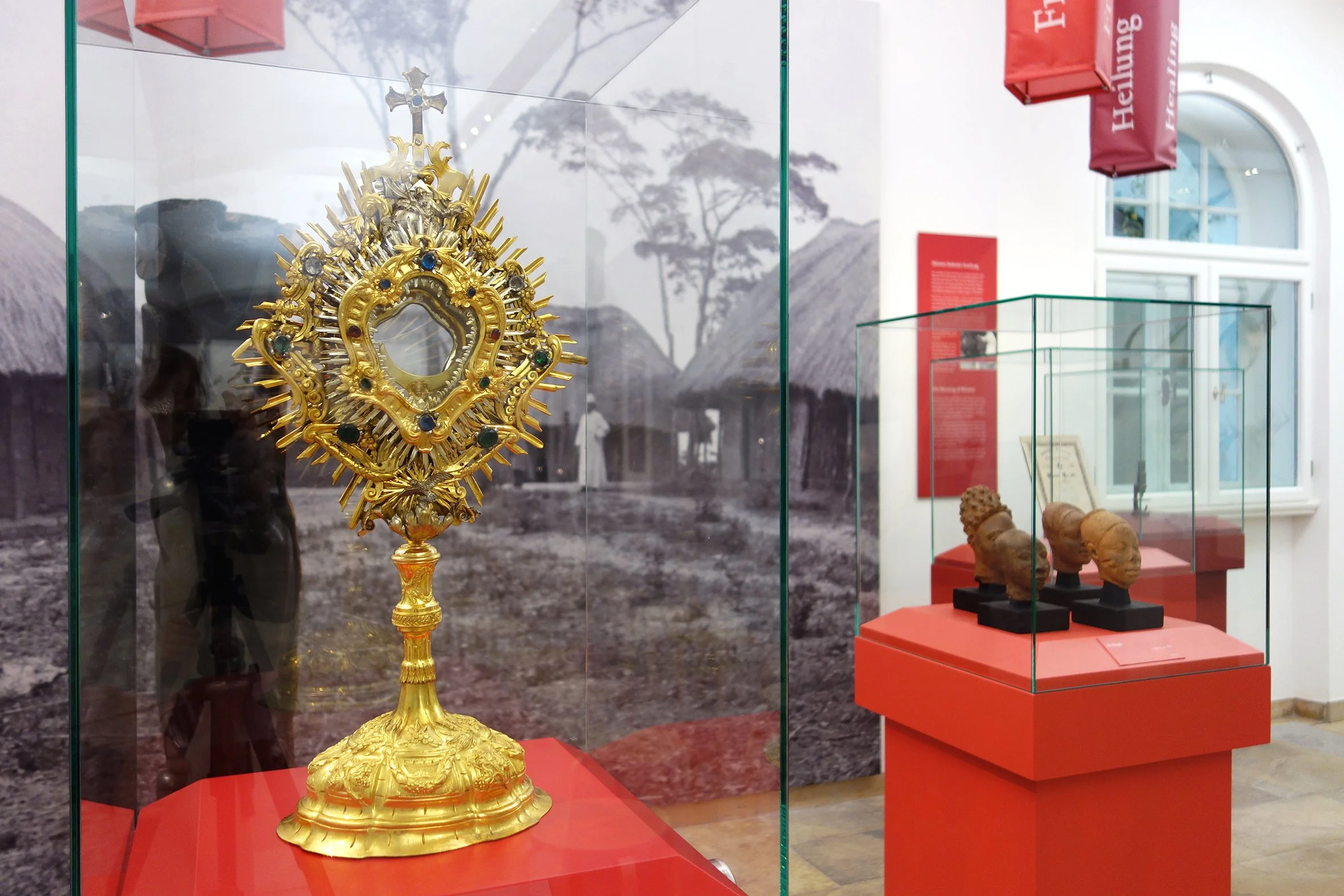 Goldenes religiöses Artefakt in einem Museum, umgeben von anderen kunstvollen Objekten, mit Hintergrund einer historischen Szene im Schwarzweiß.
