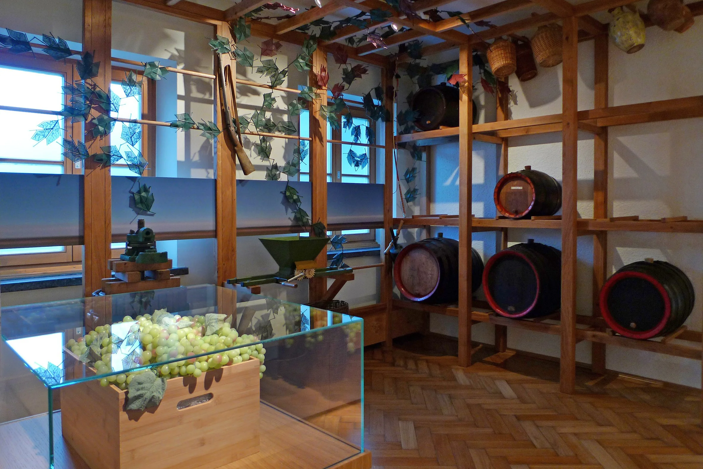 Ein Holzregal mit Weinfässern, kleiner Weinpresse und Flaschen, dekoriert mit künstlichen Efeuranken, in einem Raum mit Holzboden und Fenstern.