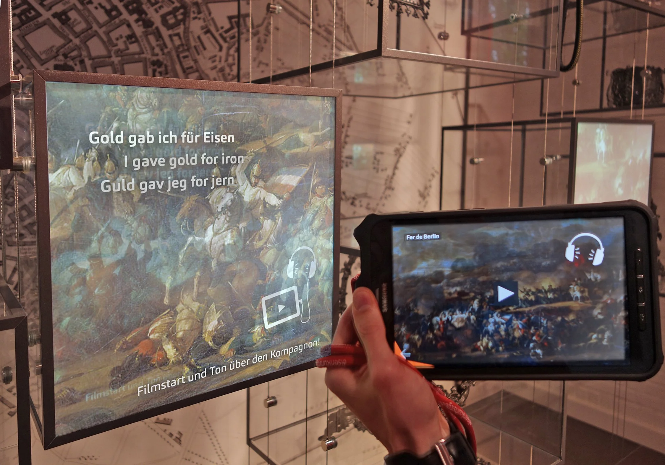 Ausstellung mit interaktivem Bildschirm, der das Gemälde 'Der Eroberer' zeigt, und eine Person, die ein Smartphone hält, um das Gemälde zu betrachten. Der Bildschirm enthält deutsche Übersetzungen des Liedtextes 'Gold gab ich für Eisen'.