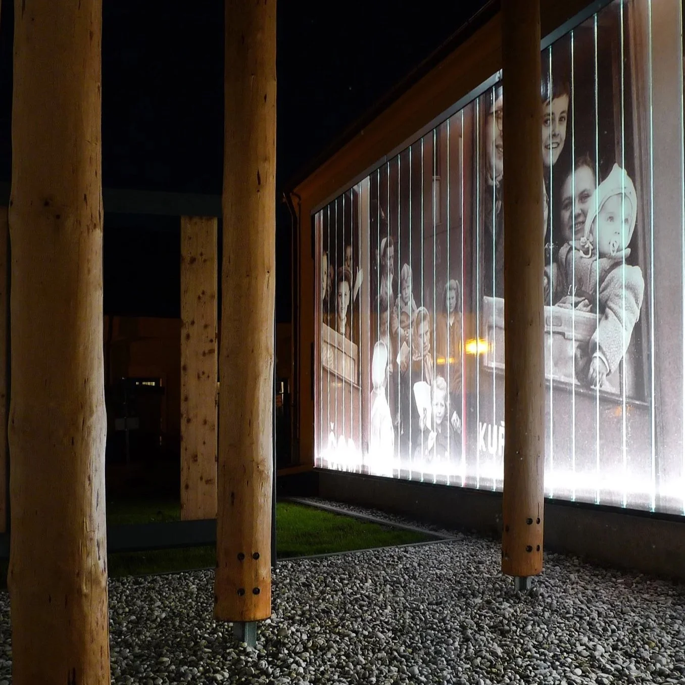 Ein beleuchtetes Bild an einer Wand mit mehreren Menschen, die auf einer Fahrt mit einem Zug oder Bus sind, im Hintergrund. Holzpfosten umgeben die Szenerie und der Boden ist mit kleinen Steinen bedeckt.