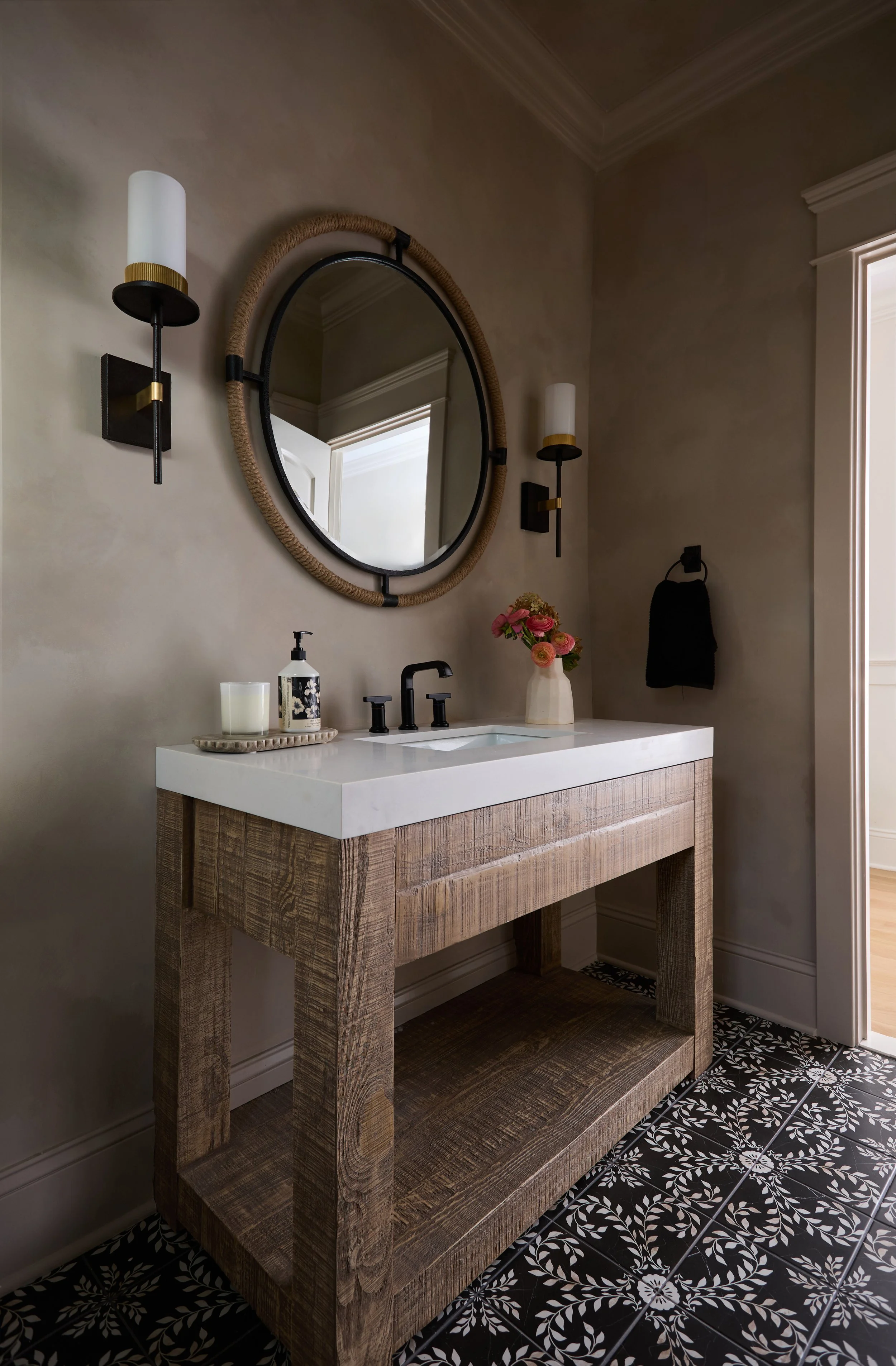 Bathroom powder room vanity with a white marble countertop, black fixtures, a round mirror with a rope frame, black and gold wall sconces with white shades. The floor has black and white patterned tiles.