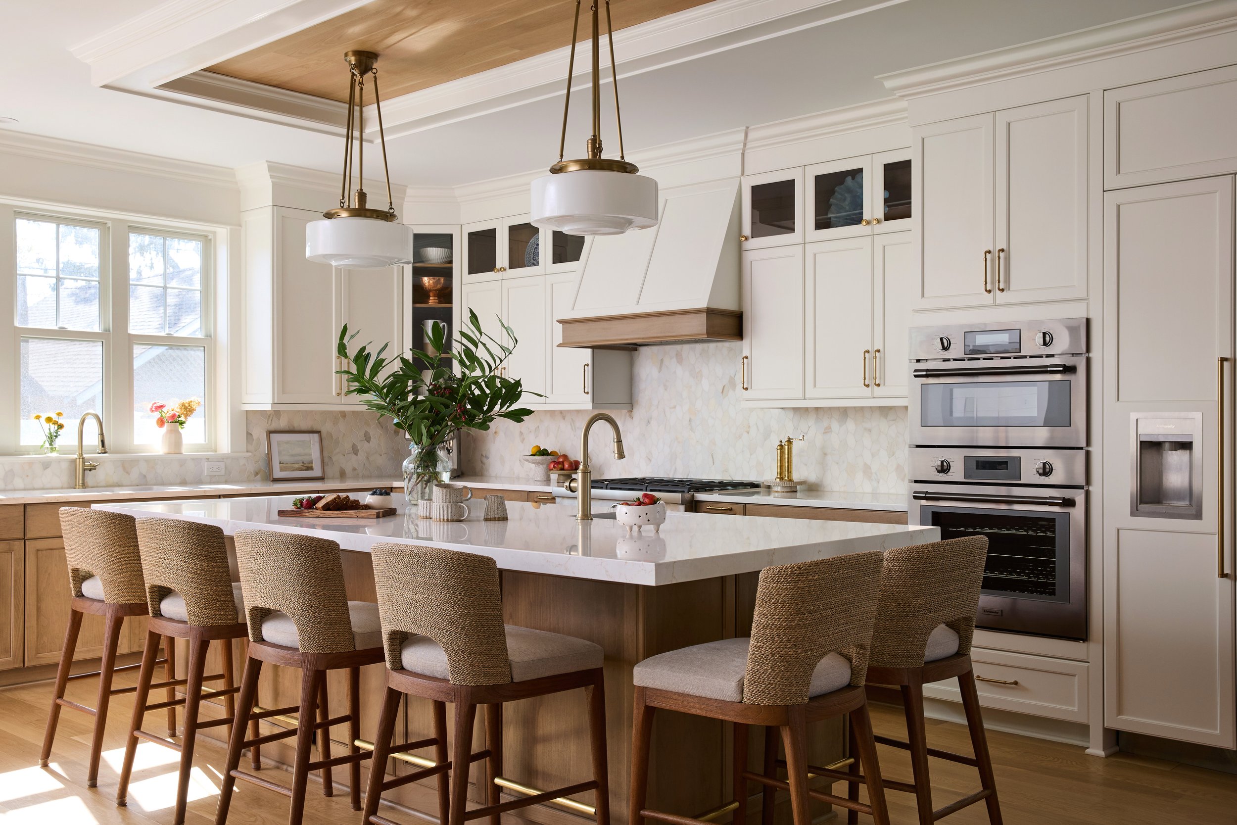 Modern coastal kitchen with white upper cabinets, warm wood lower cabinets, a large island with upholstered seagrass stools, stainless steel appliances, large pendant lighting, illuminated by natural light from large windows.