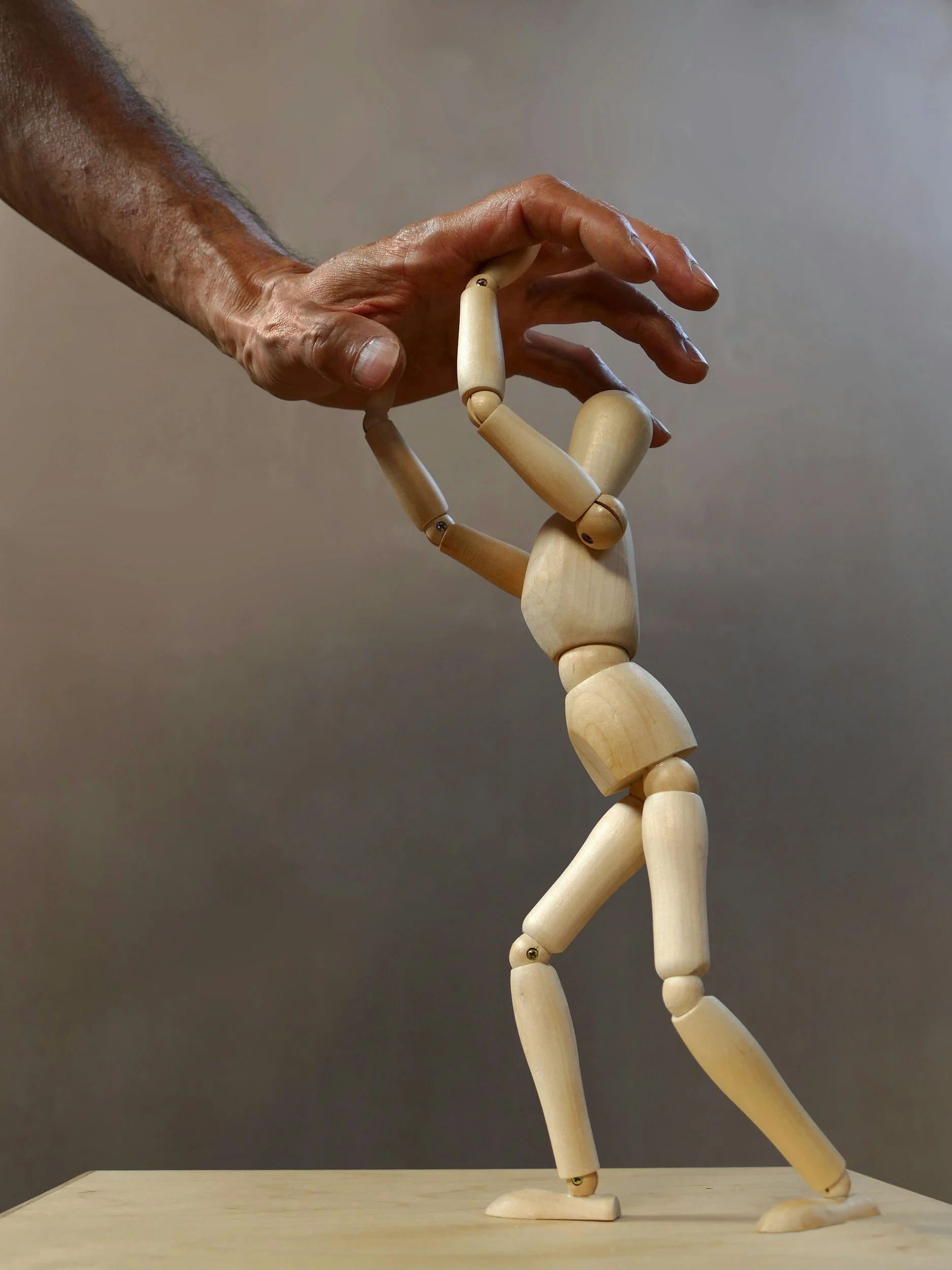 A wooden artist mannequin posed on a flat surface, with a human hand gently touching its head. The mannequin's left arm is bent, and its right arm is extended downward. The background is plain and neutral.