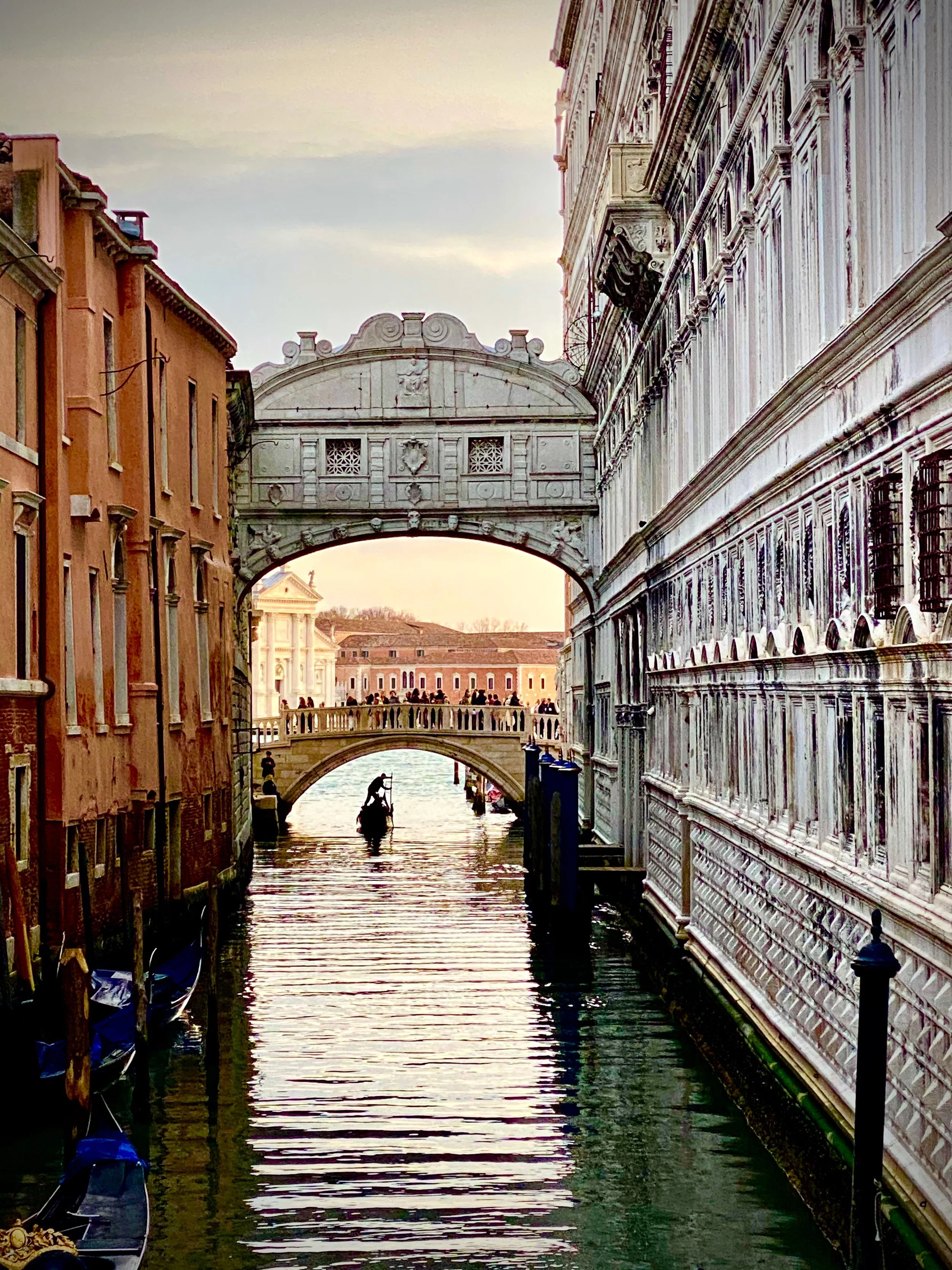 View of a canal in Venice, Italy, with historic buildings on both sides and a stone bridge in the distance. Gondolas are docked along the edges, and a gondola with a passenger is in the water.