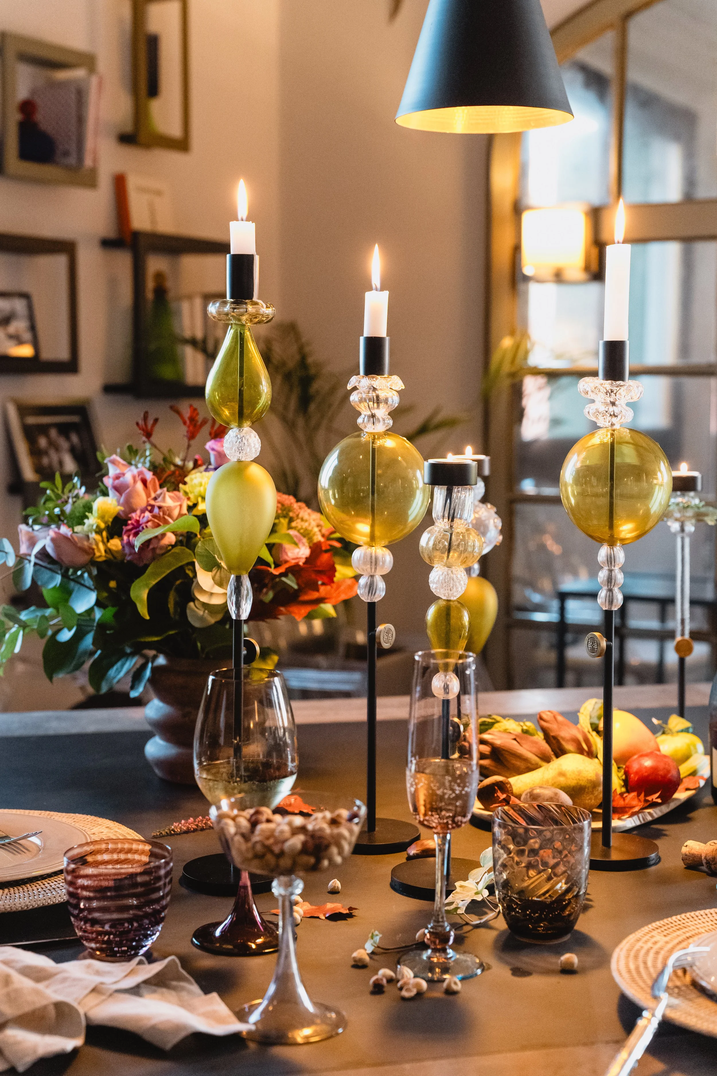 Decorative table centerpiece with three lit candles in black and gold candle holders, surrounded by a floral arrangement, glassware, and a fruit platter on a dinner table in a warmly lit room.