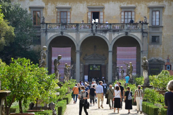 Tourists walking through a historic building's garden with statues, leading to a grand entrance with an arched portico, above which is a balcony with windows and a yellow facade.