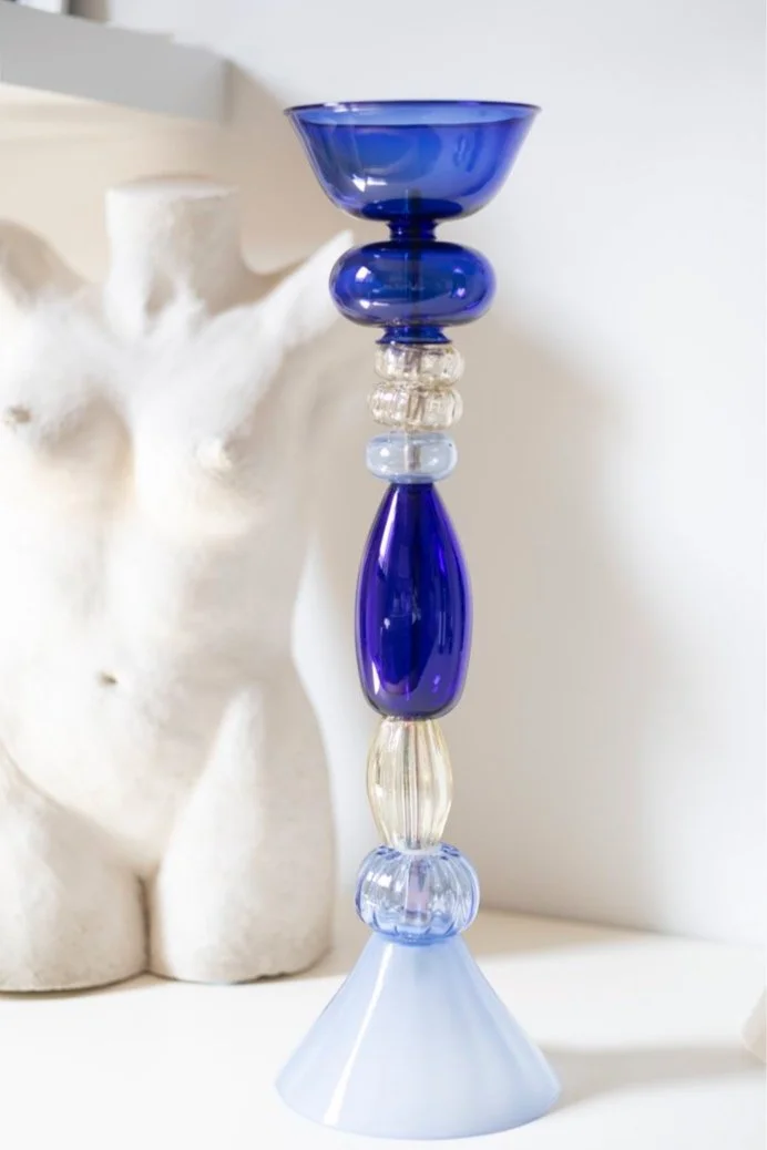 A tall decorative glass candlestick holder with blue, clear, and lavender glass sections on a white surface, with white decorative sculpture in the background.