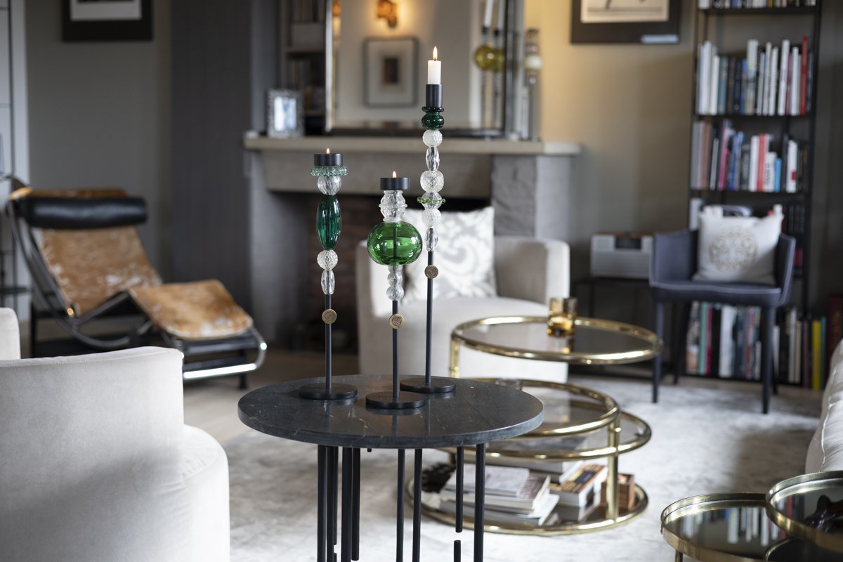 Living room with a black marble side table holding three tall candlestick holders with candles, surrounded by modern furniture, a white couch, a black armchair, a gold nested coffee table, and a tall bookcase filled with books.