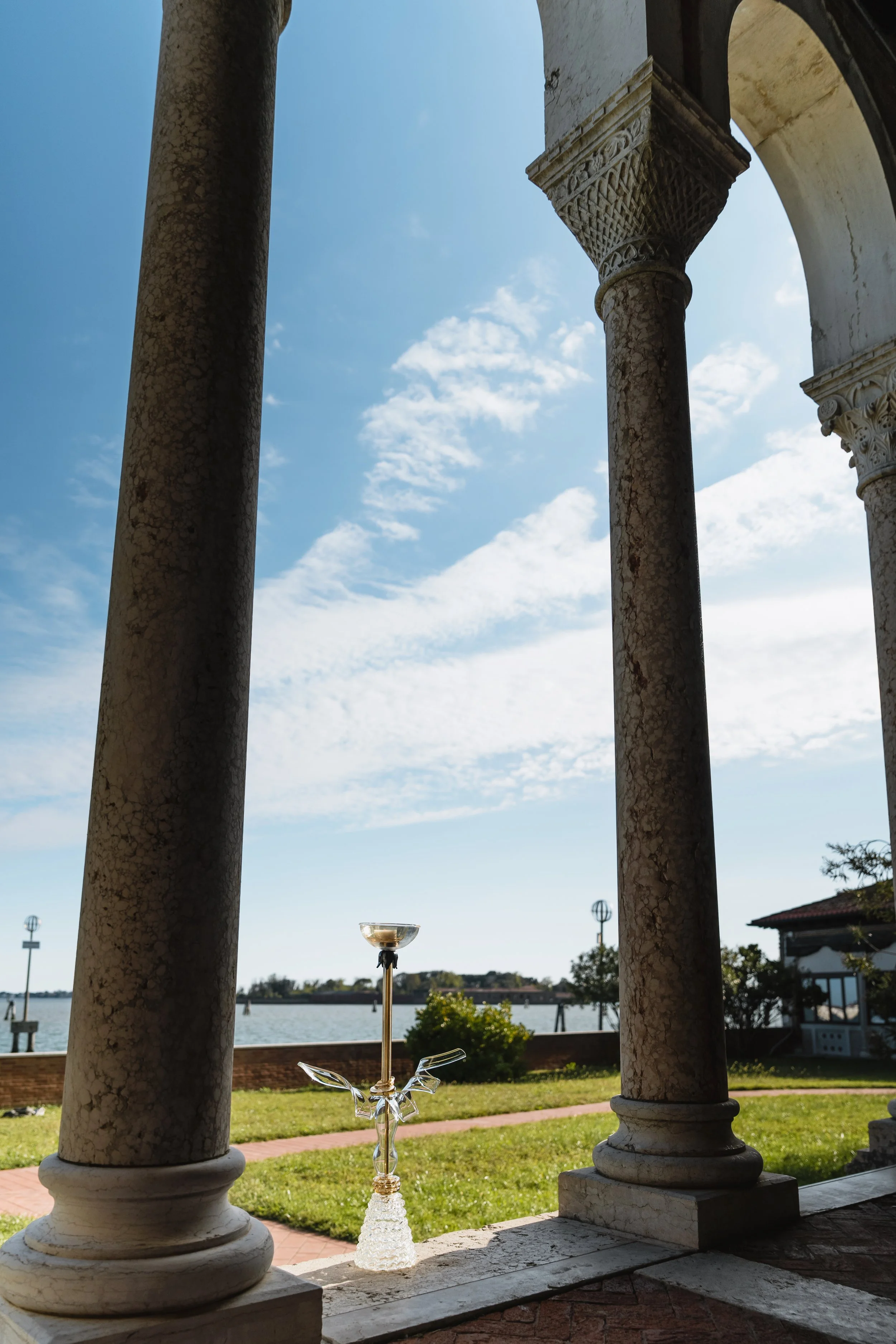 View from a stone balcony with ornate columns overlooking a river, with a glass sculpture of a dragon and a glass vessel on top of a stand in the foreground, a clear blue sky with clouds, and a building with a red roof in the background.